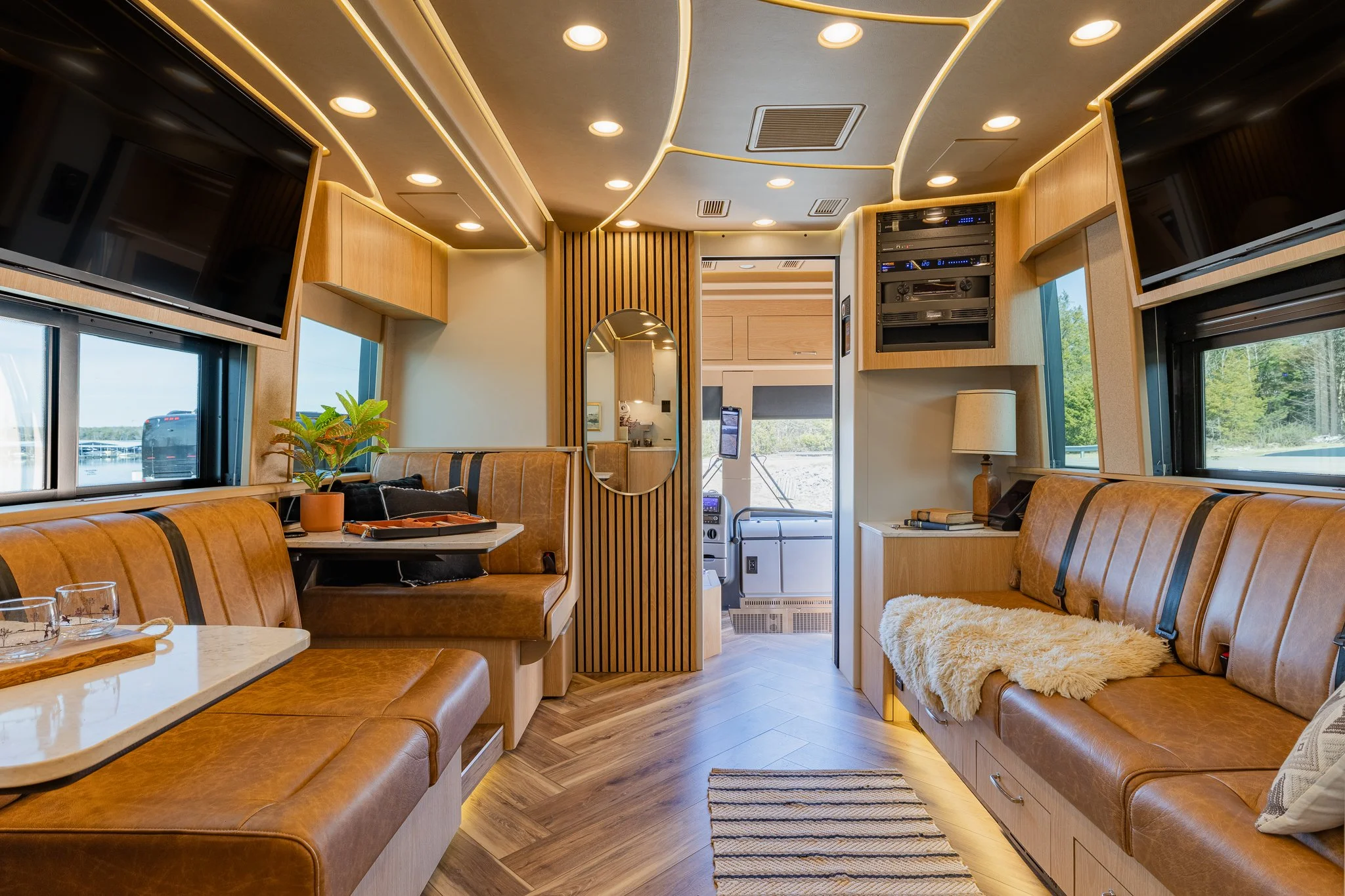 Interior of a modern RV with leather seating, wood accents, and multiple windows showing a scenic outdoor view.