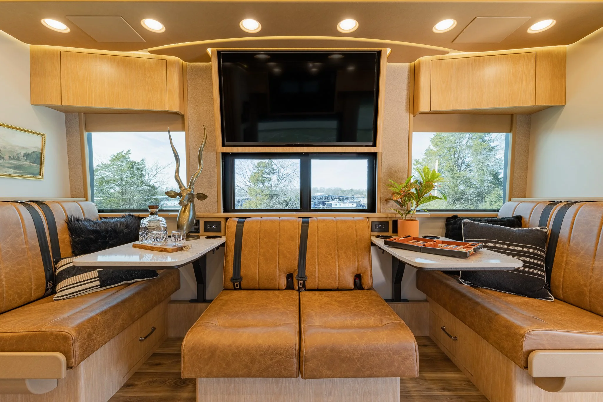 A cozy indoor space with two cushioned benches on either side of a window, a TV mounted above a central window, decorated with a plant, a decorative deer sculpture, and glassware, with warm lighting and wood accents.