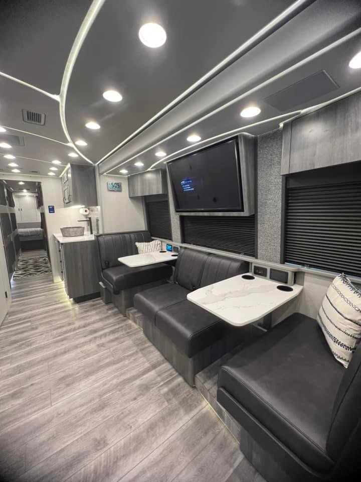 Interior of a modern bus or RV with black leather seating, white tables, a flat-screen TV, and black window coverings.