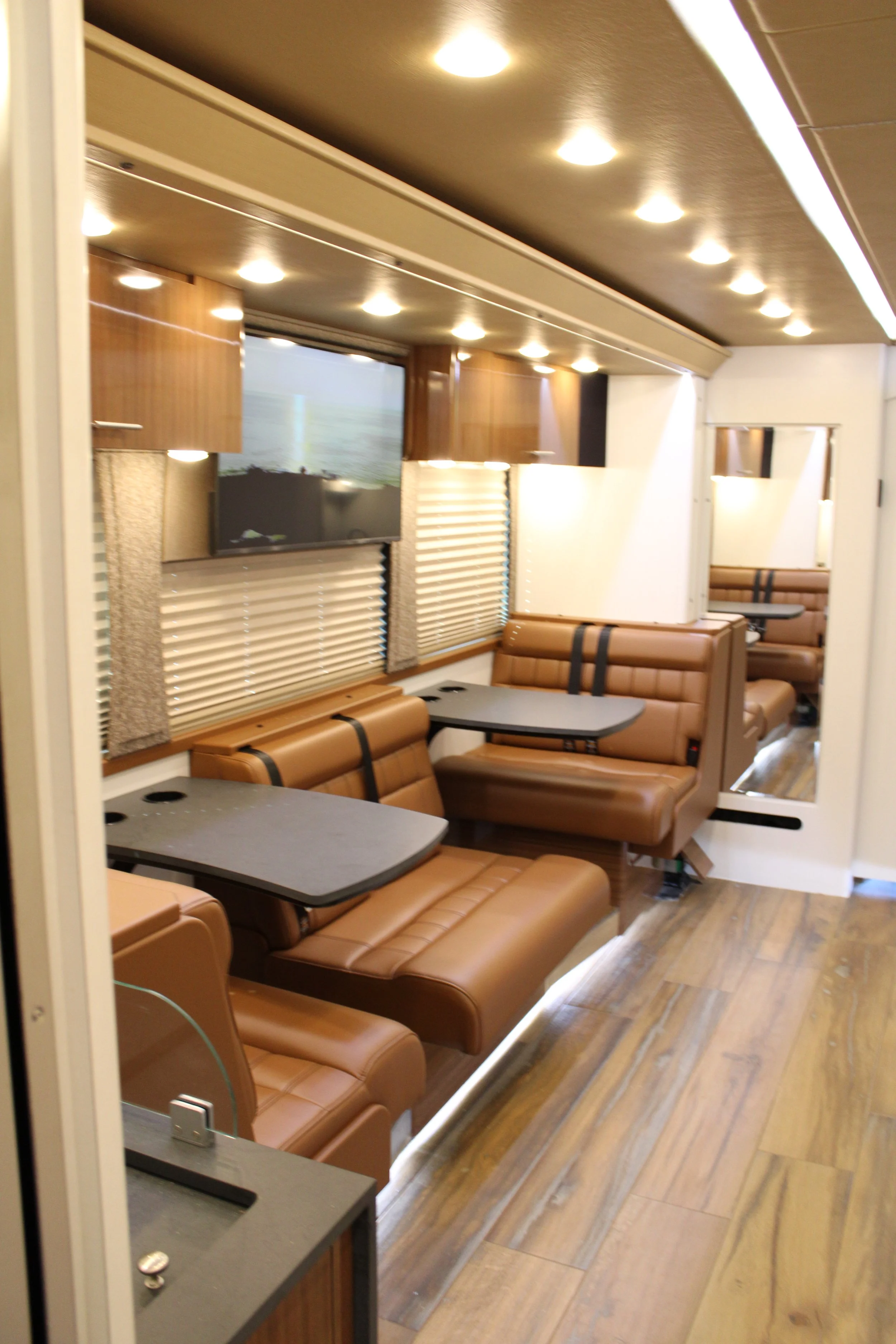 Interior of a bus or train with leather seating, foldable tables, wooden cabinets, a TV, and blinds on the windows.