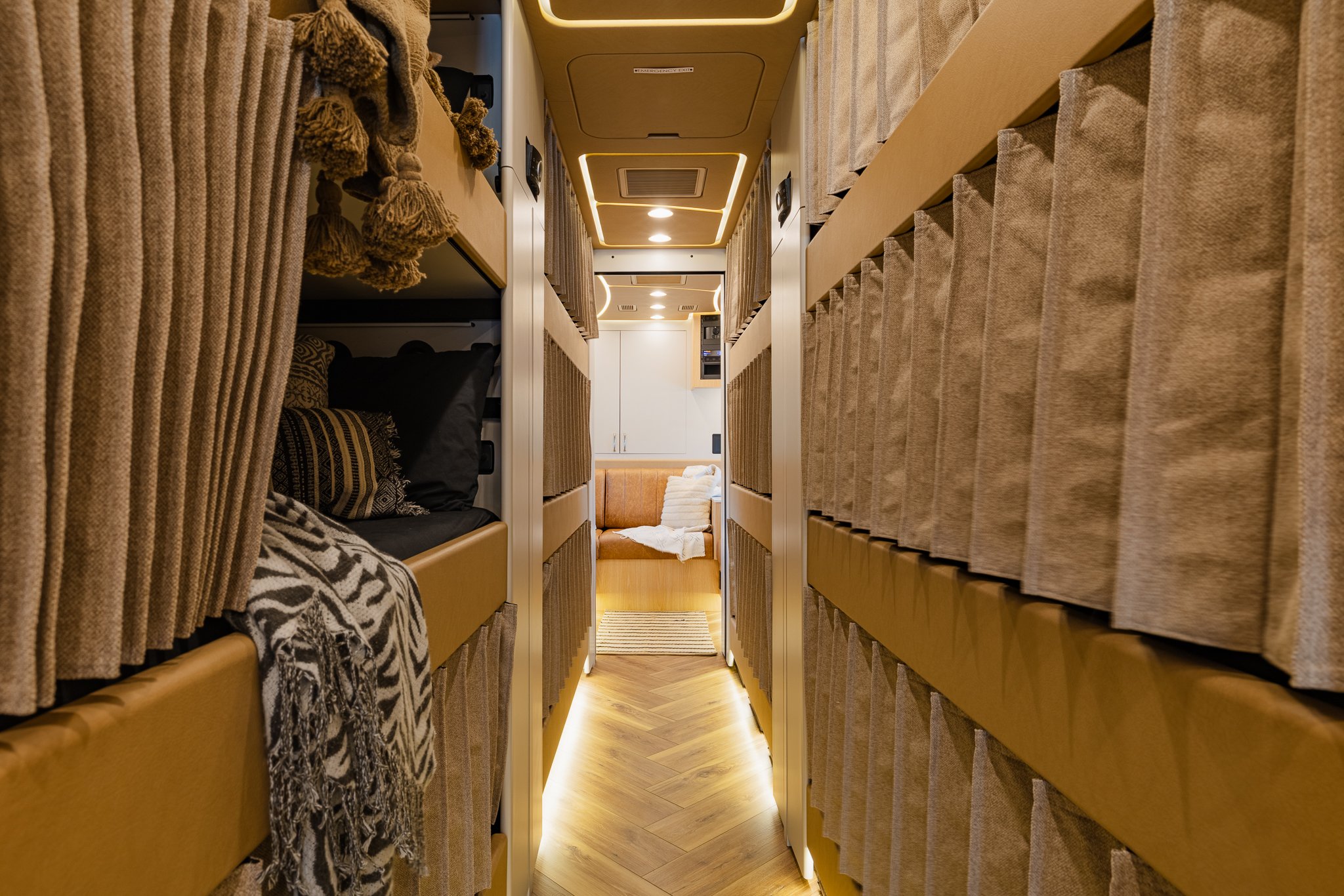 Inside a luxury bus or train with beige and brown interior, including shelves with storage, curtains, and seating area with blankets and pillows at the end of the corridor.
