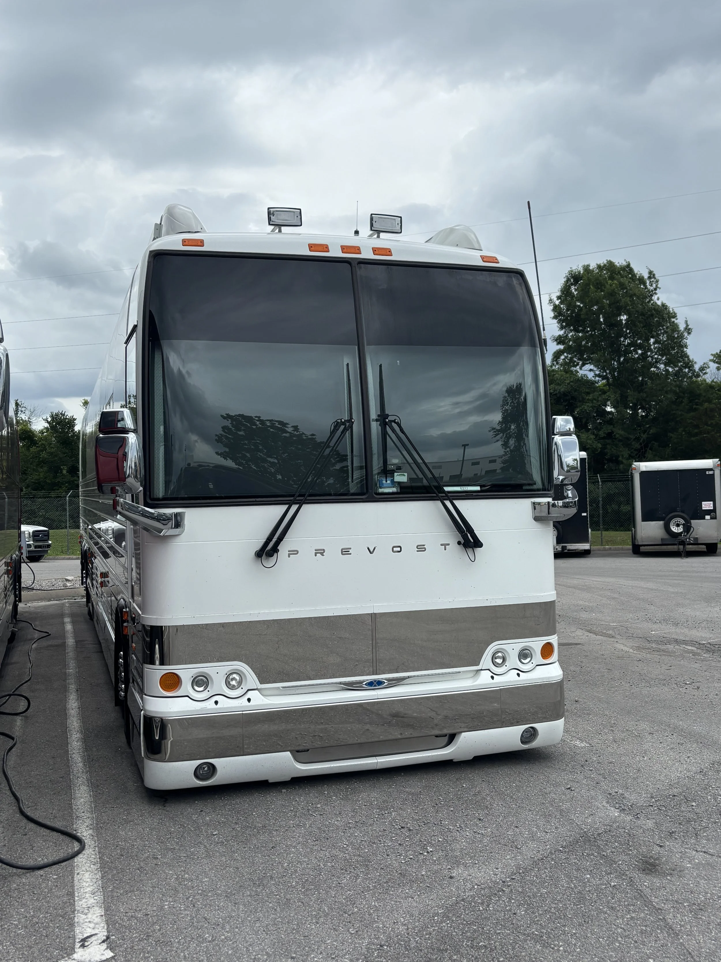 Front view of a white Prevost bus parked in a lot, with trees and other vehicles in the background under a cloudy sky.