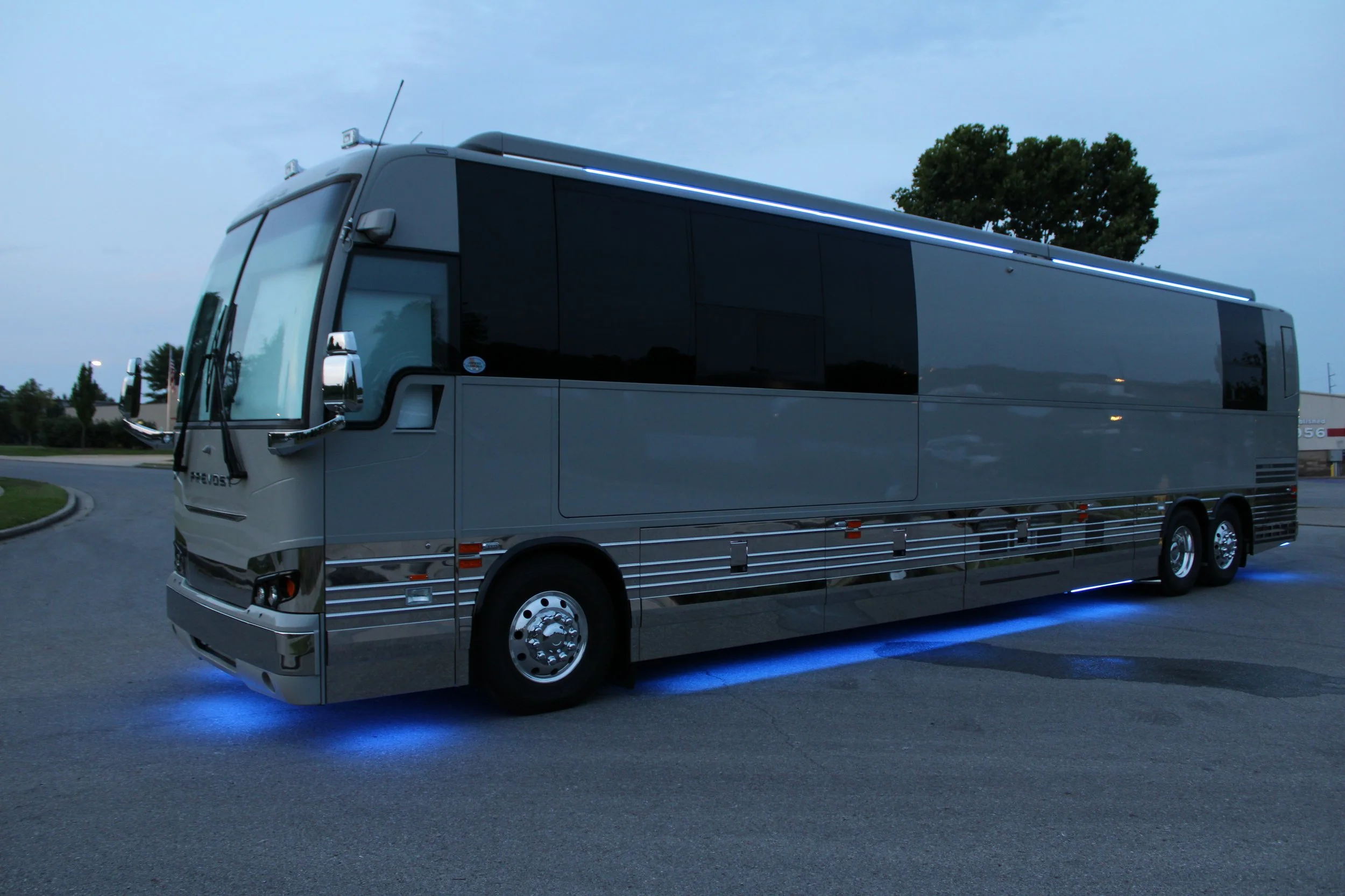 A large, modern, silver bus with blue underglow lighting parked on a paved area during dusk or early evening, with trees and a building in the background.