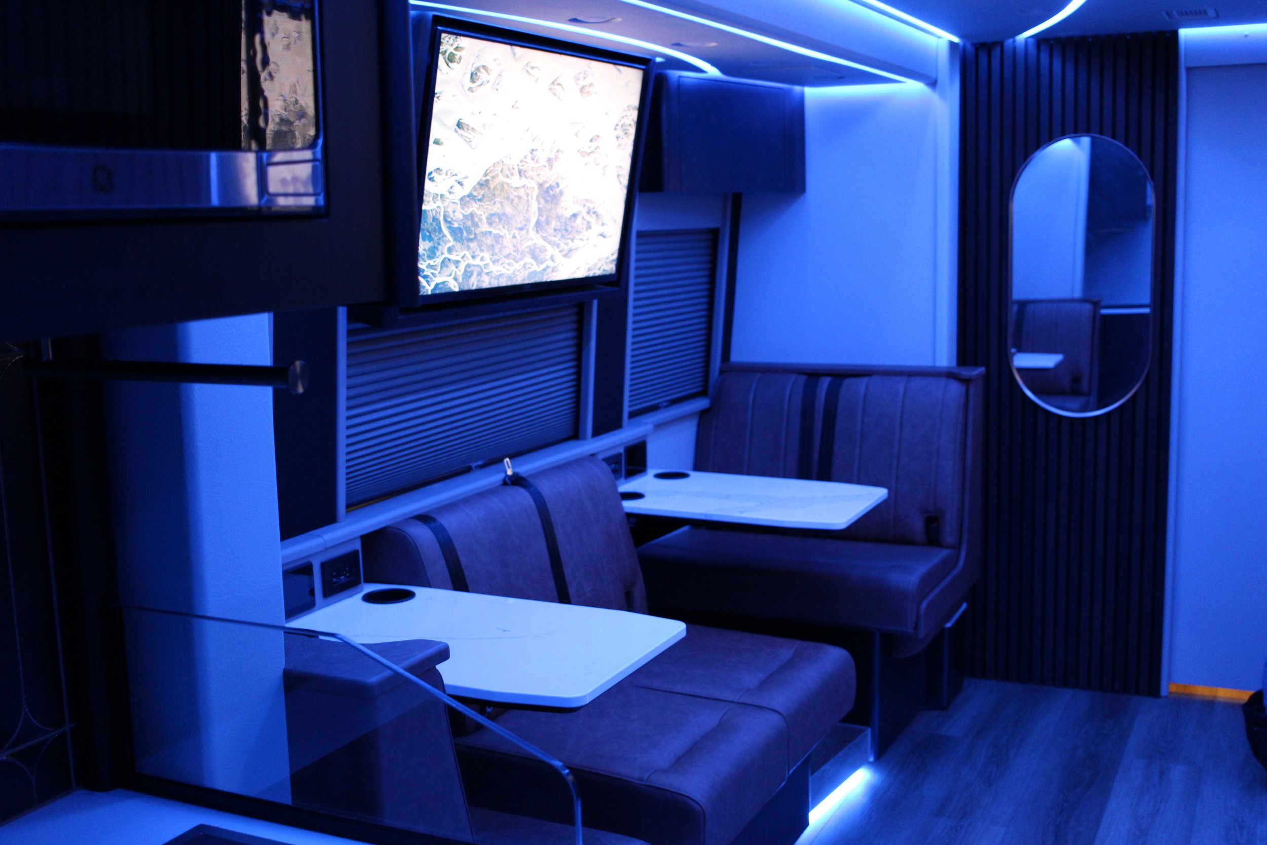A room with blue lighting, featuring two wall-mounted TVs, a booth-style seating area with a table, and a mirror on the wall.