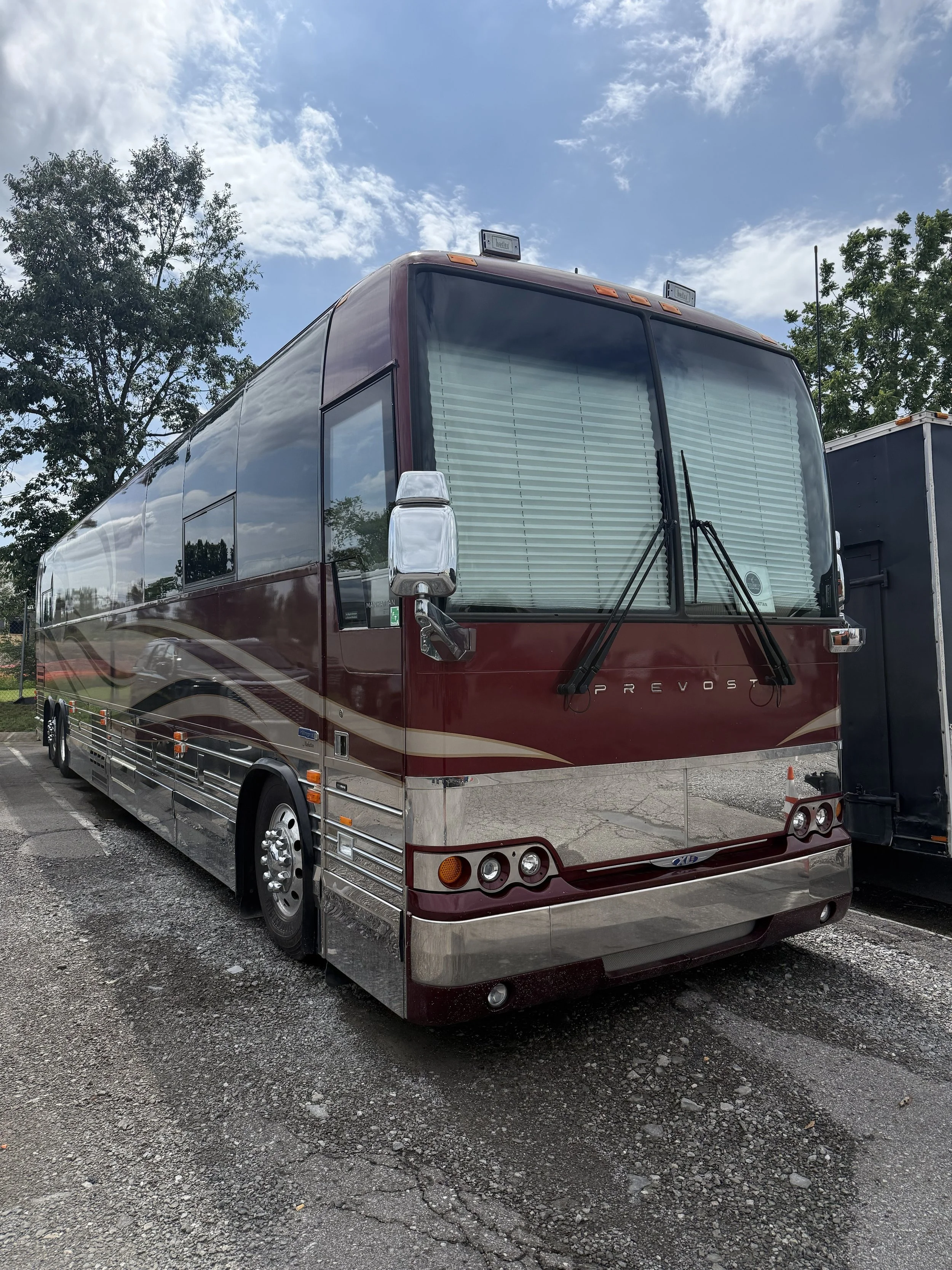 A maroon and chrome bus parked in a lot with trees and a partly cloudy sky in the background.
