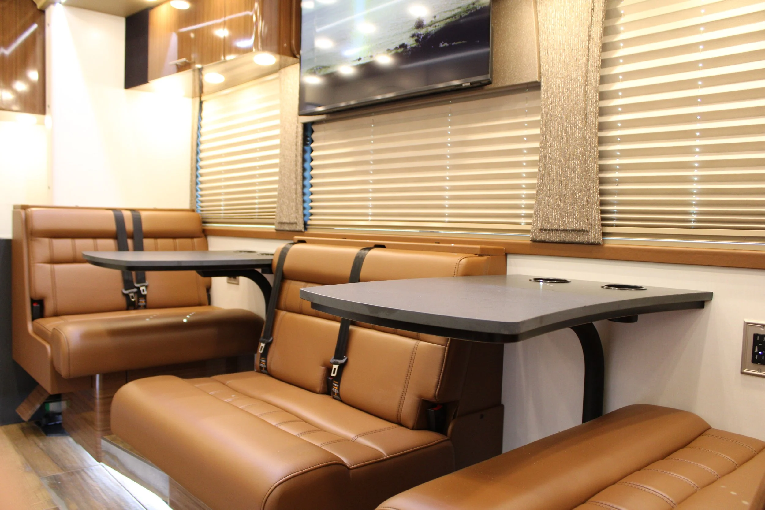 Inside a bus or train with brown leather seats, gray tables, window blinds, and a mounted television screen.
