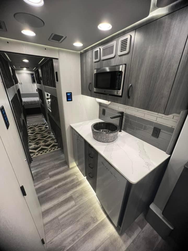 Small modern kitchen with gray cabinetry, white marble countertop, stone vessel sink, black faucet, microwave, gray tile backsplash, and wood-look flooring. A hallway with bunk beds is visible in the background.