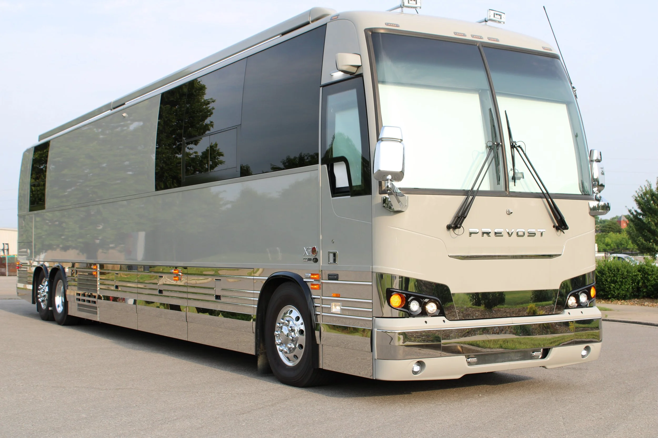 A shiny gray luxury bus parked on a paved road with trees and a cloudy sky in the background.