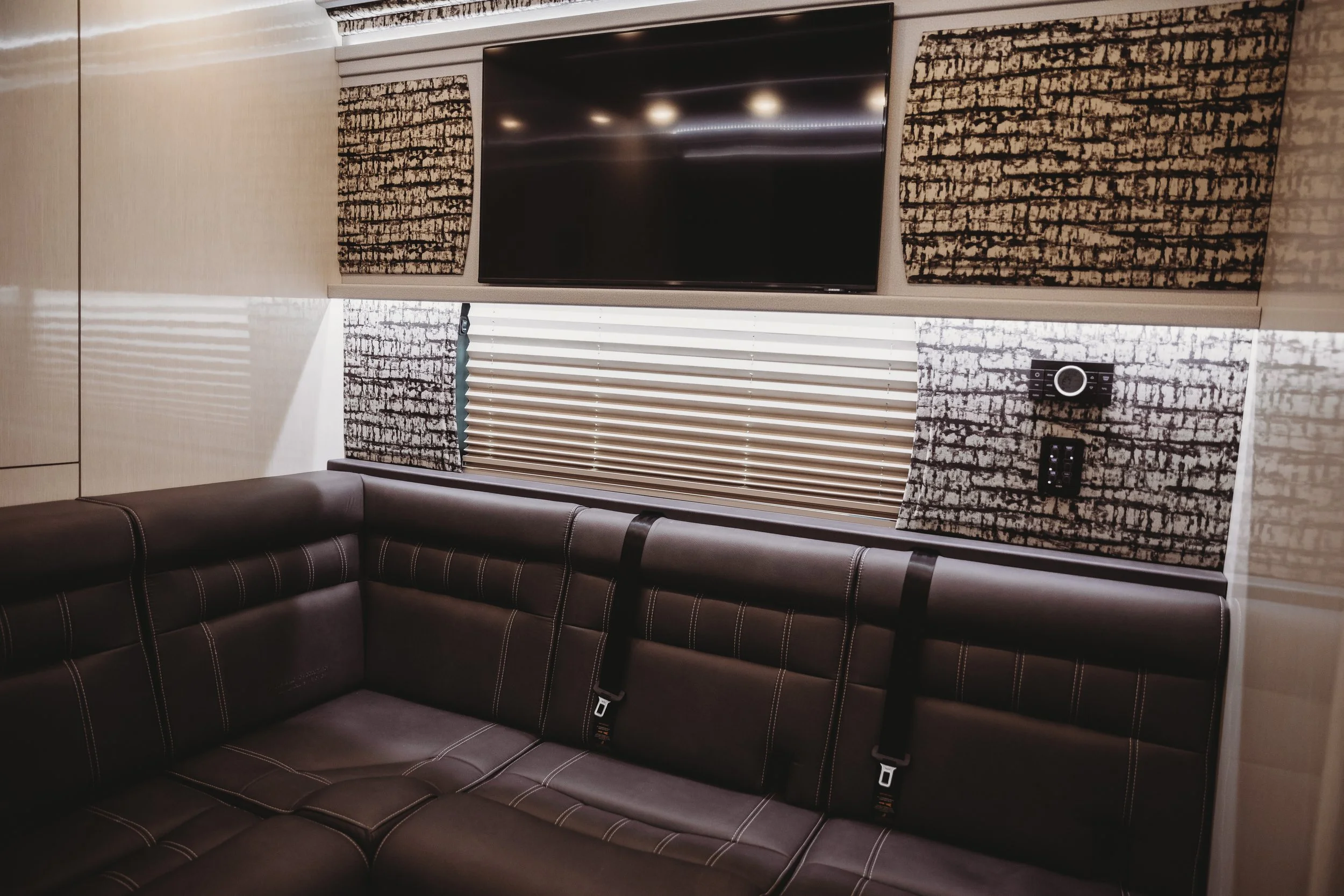 Indoor space with a black leather sectional sofa, a wall-mounted television, window with blinds, wall-mounted control panel, and patterned wall panels.