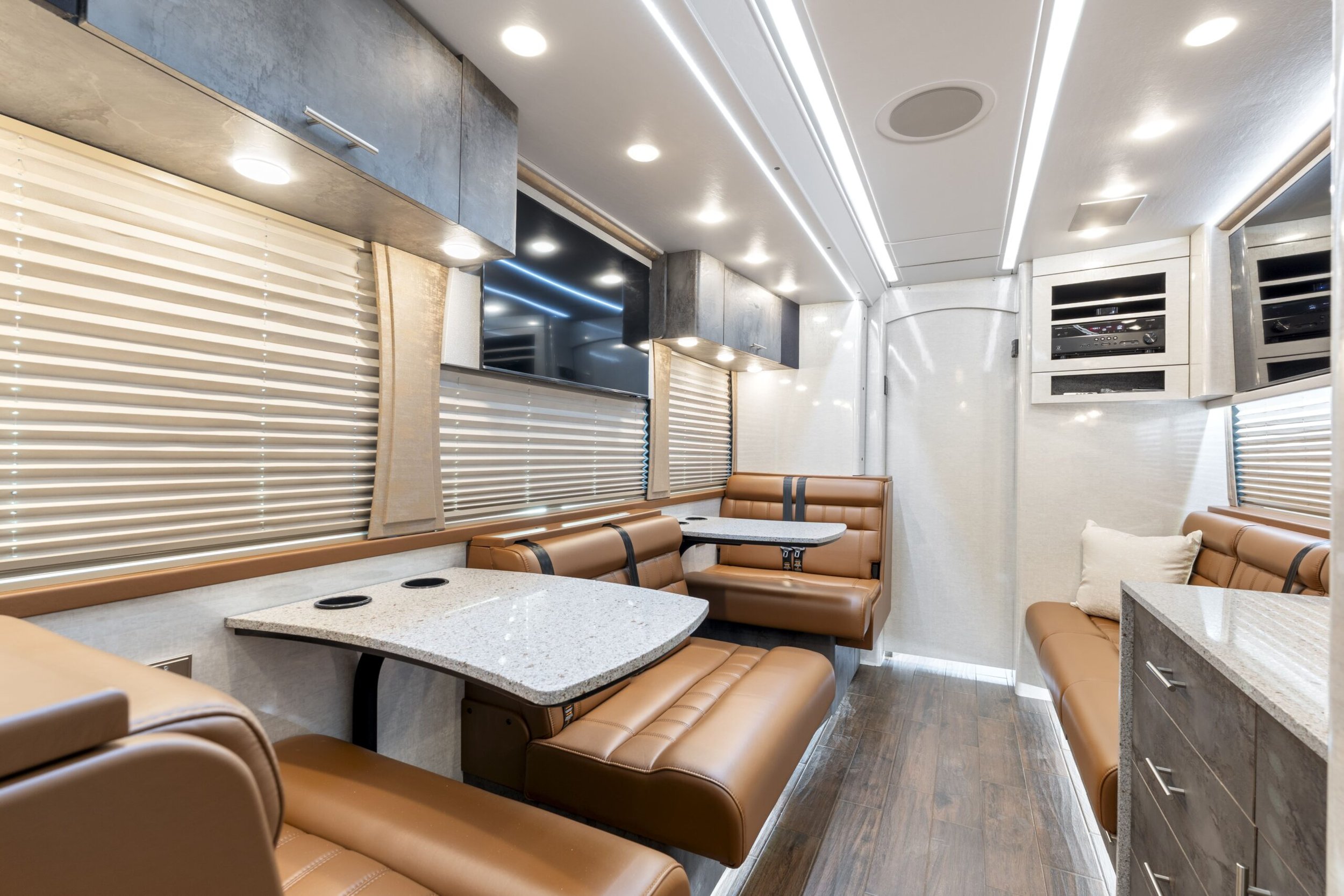 Interior of a modern recreational vehicle or bus, featuring tan leather seats, granite countertops, and windows with beige blinds.