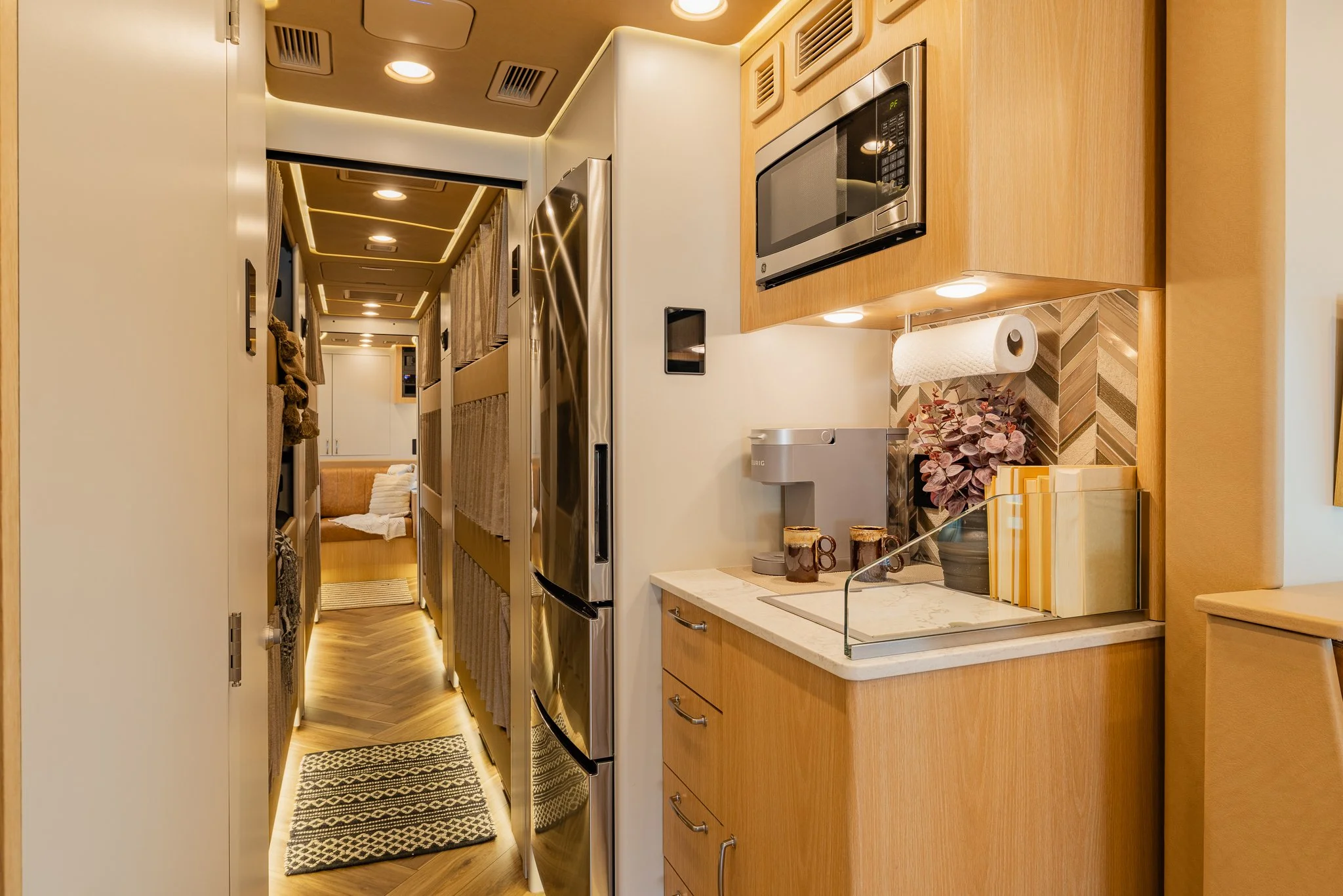 Interior of a tiny kitchen with wooden cabinets, stainless steel refrigerator, microwave, and a coffee station with paper towels and decorative plant, leading into a cozy living area with a brown sofa.
