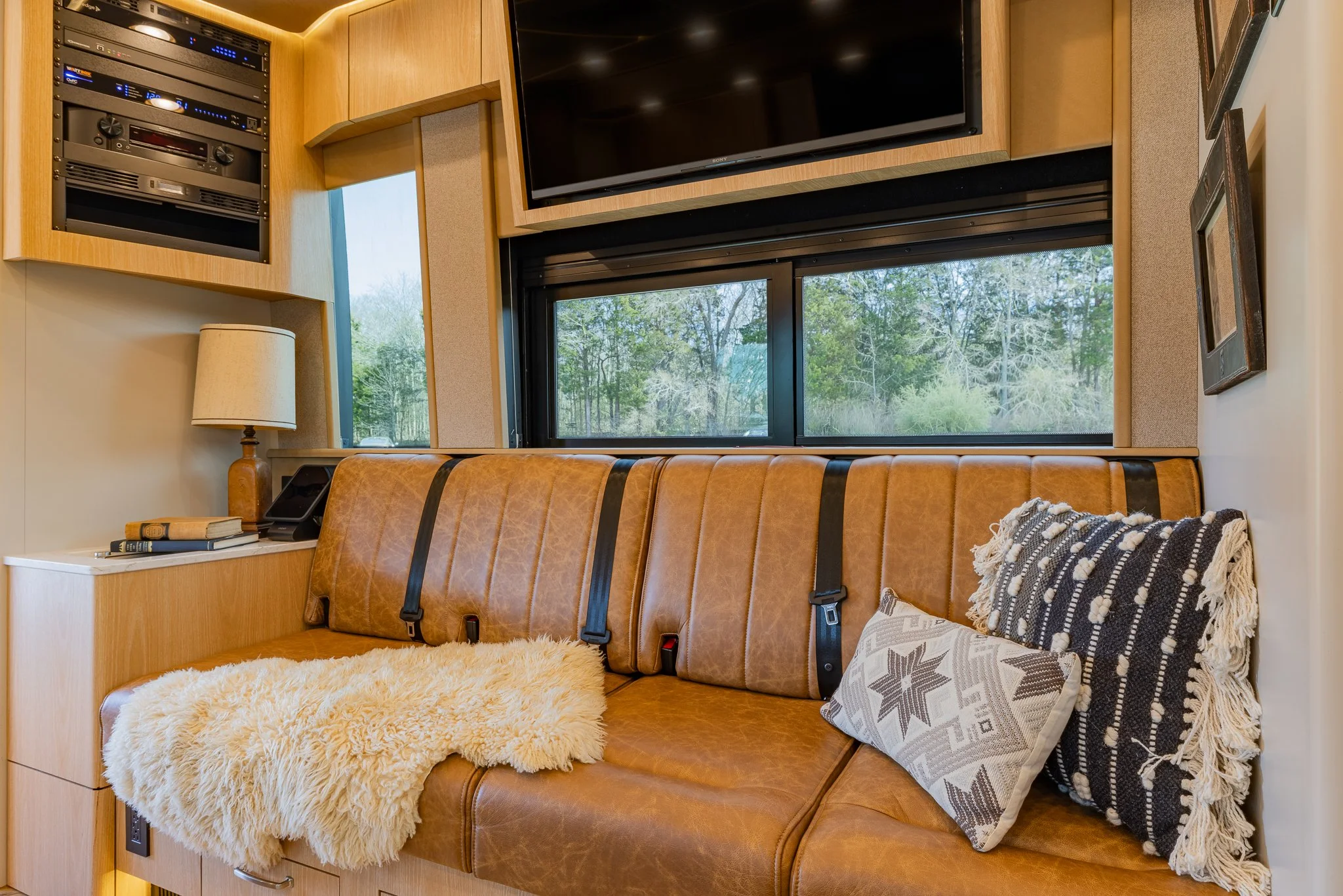 Interior of a cozy room with a leather couch, decorative pillows, a blanket, a small lamp, and a large window showing trees outside. Wall-mounted TV and audio equipment are also visible.