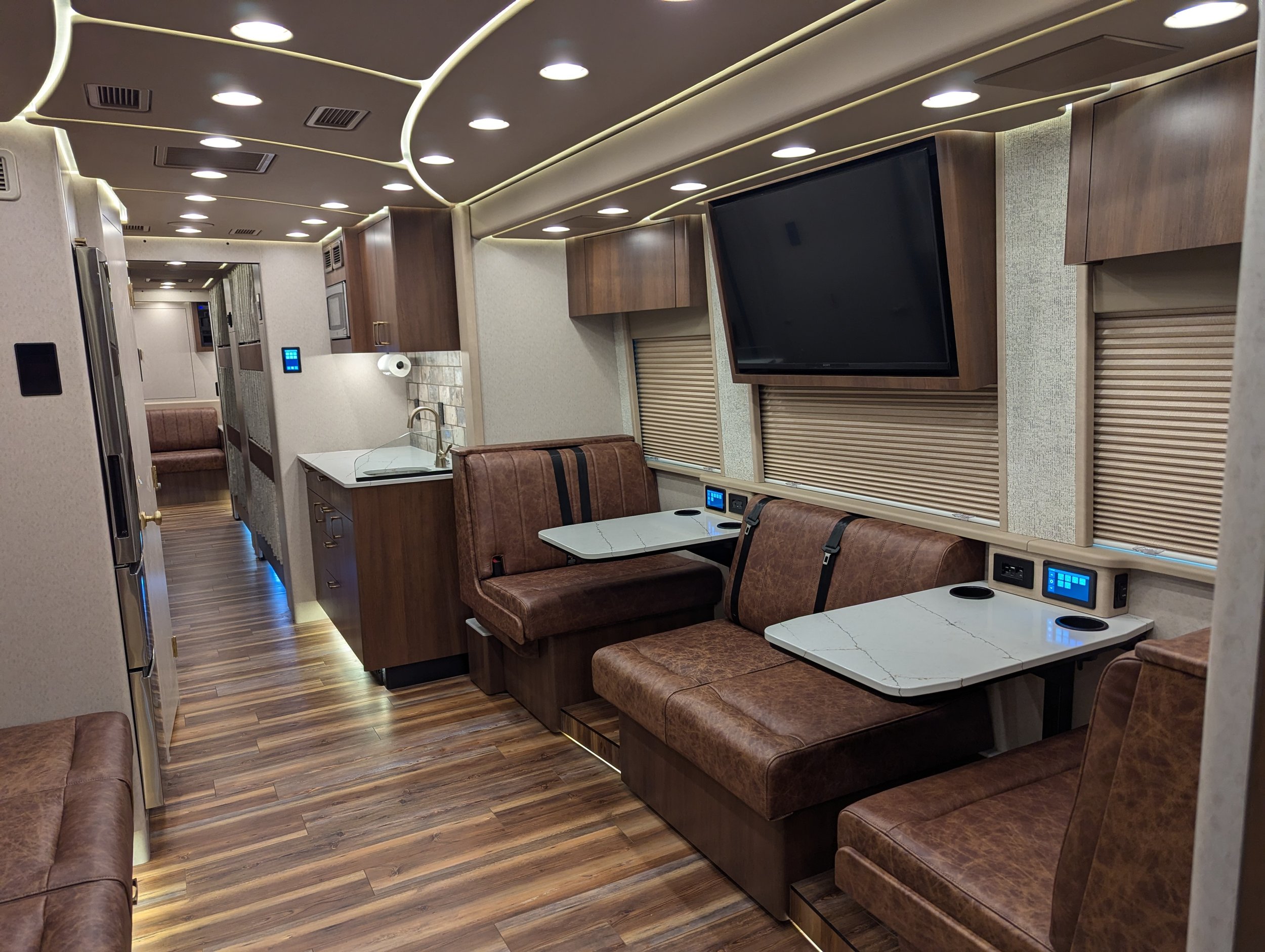 Interior of a modern RV with brown leather seating, white marble-style tables, wood floors, a large flat-screen TV, and window blinds.