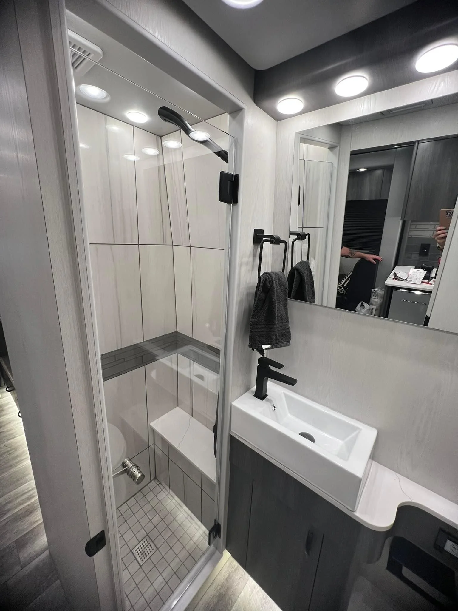 Modern bathroom with a walk-in shower, white tiles, a black handheld showerhead, a small white sink with a black faucet, and a large mirror reflecting part of the room and a person taking the photo.
