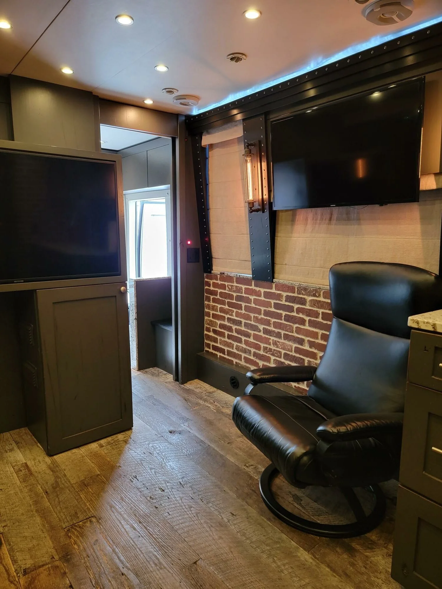 Interior of a modern office or studio with a black leather swivel chair, two large flat-screen televisions mounted on the brick and wood wall, a small overhead light fixture, and a doorway leading outside. Wood flooring and dark decor create an indus
