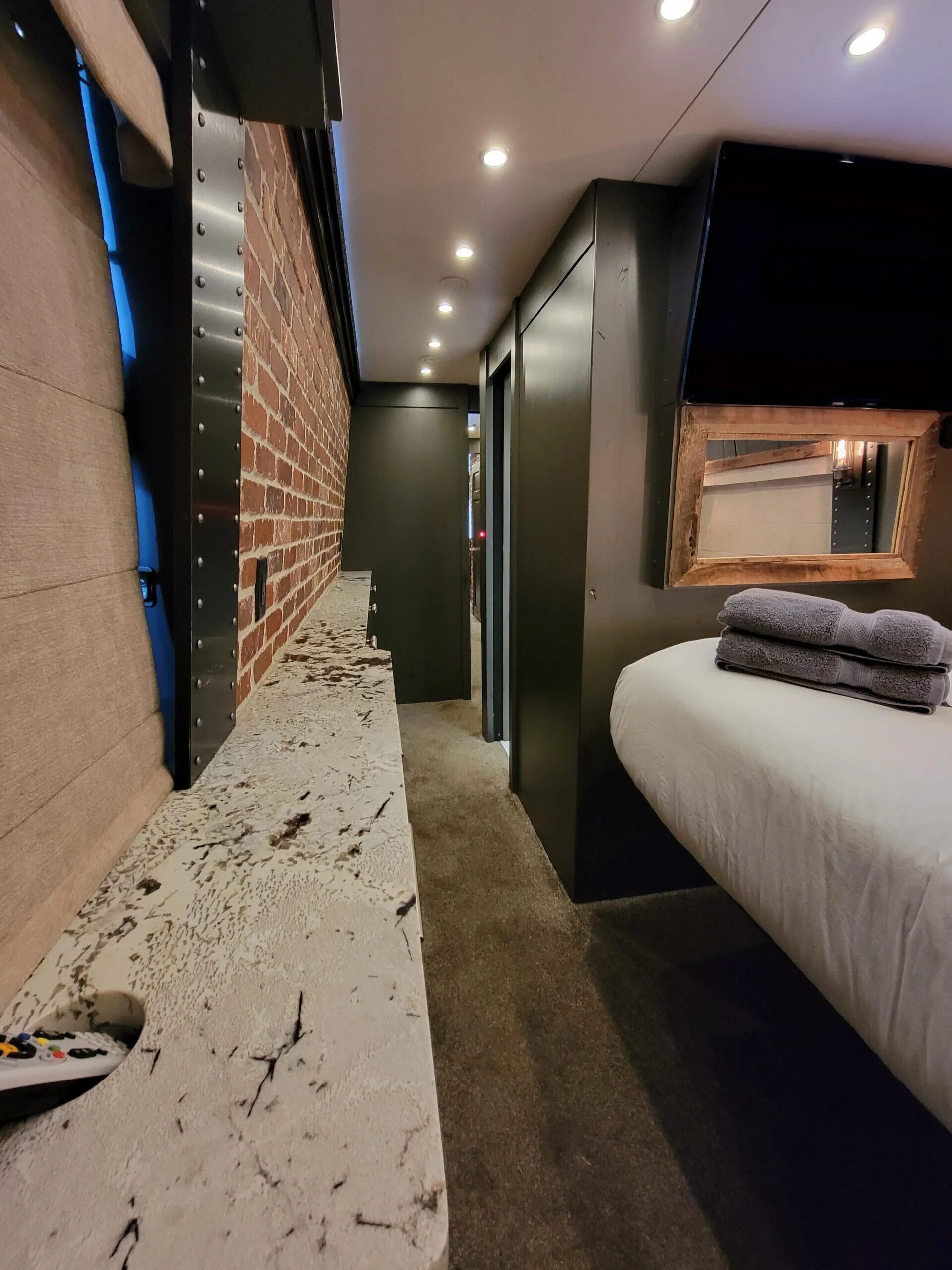 A hotel room with a bed, towels, a flat-screen TV, a mirror, a marble countertop, and a brick wall.