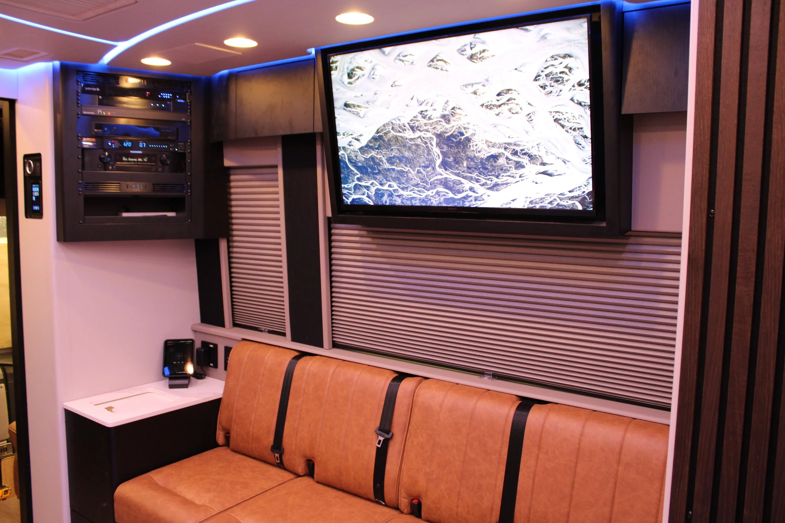 Comfortable seating area with a brown leather couch, flat-screen TV displaying a snowy mountain landscape, and an entertainment unit with electronic equipment and remote controls, in a modern interior with wood paneling and ambient lighting.