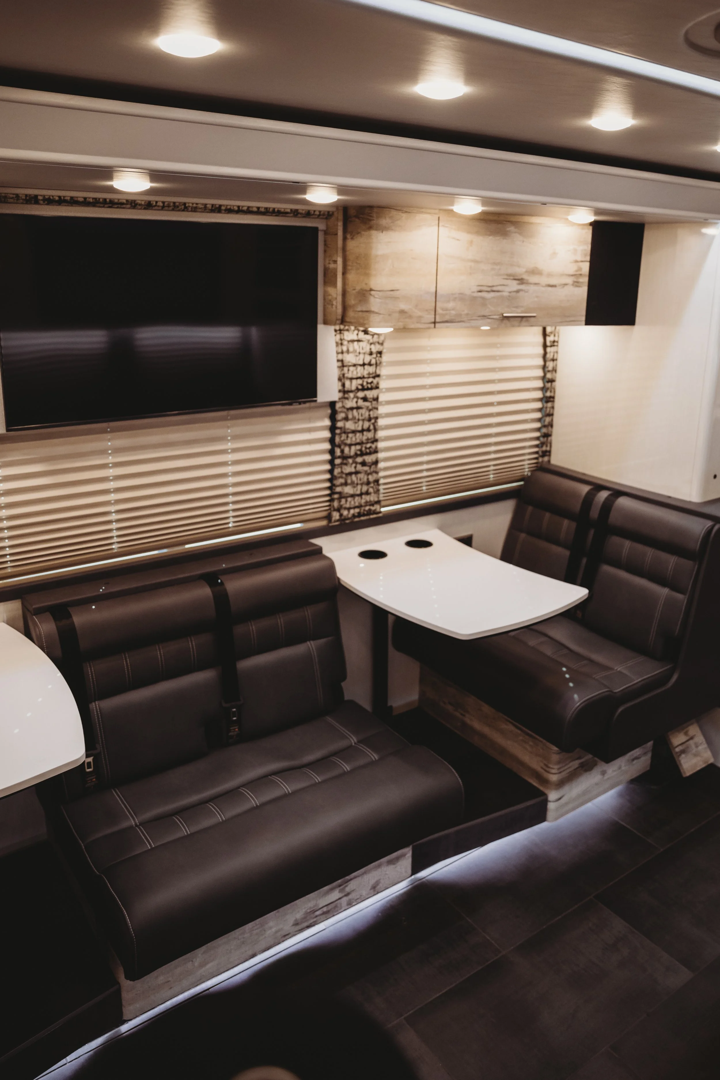 Interior of a modern RV or camper with a mounted TV, brown leather seating, and a small white table between the seats. Window blinds and ceiling lights are visible.