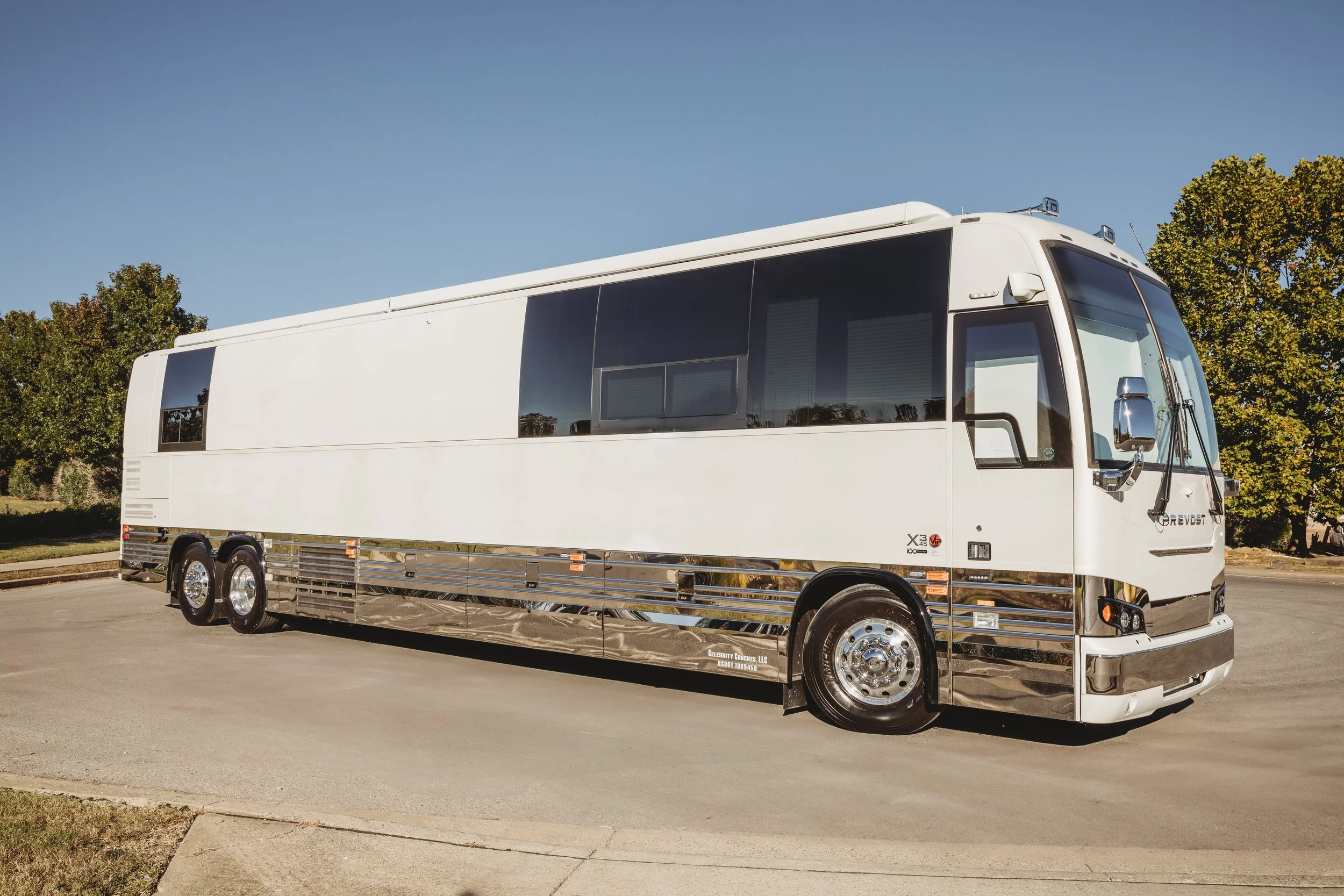 White luxury bus parked on the side of the road with clear sky and trees in the background.