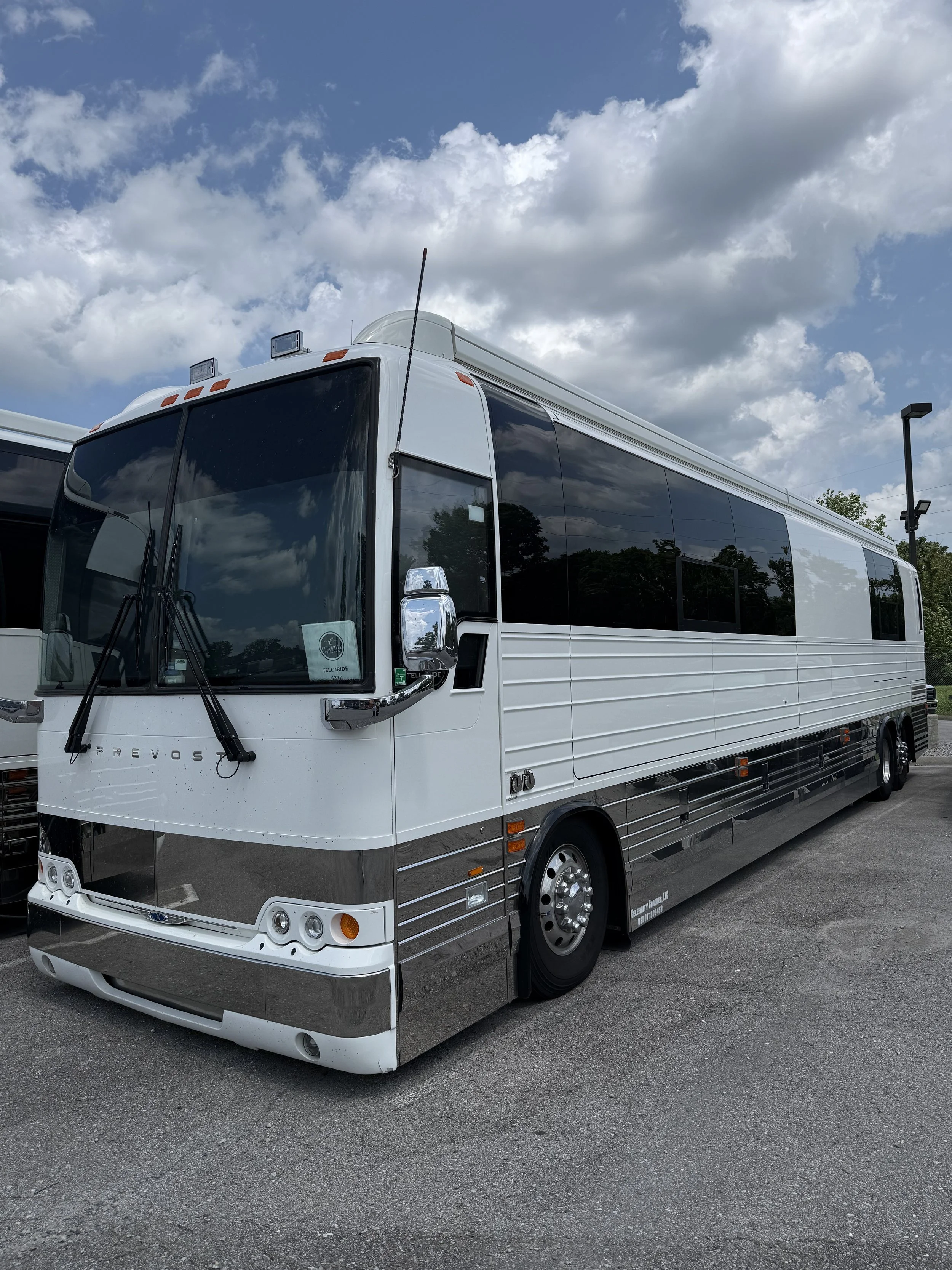 A white luxury tour bus with reflective tinted windows parked on a paved lot under a partly cloudy sky.