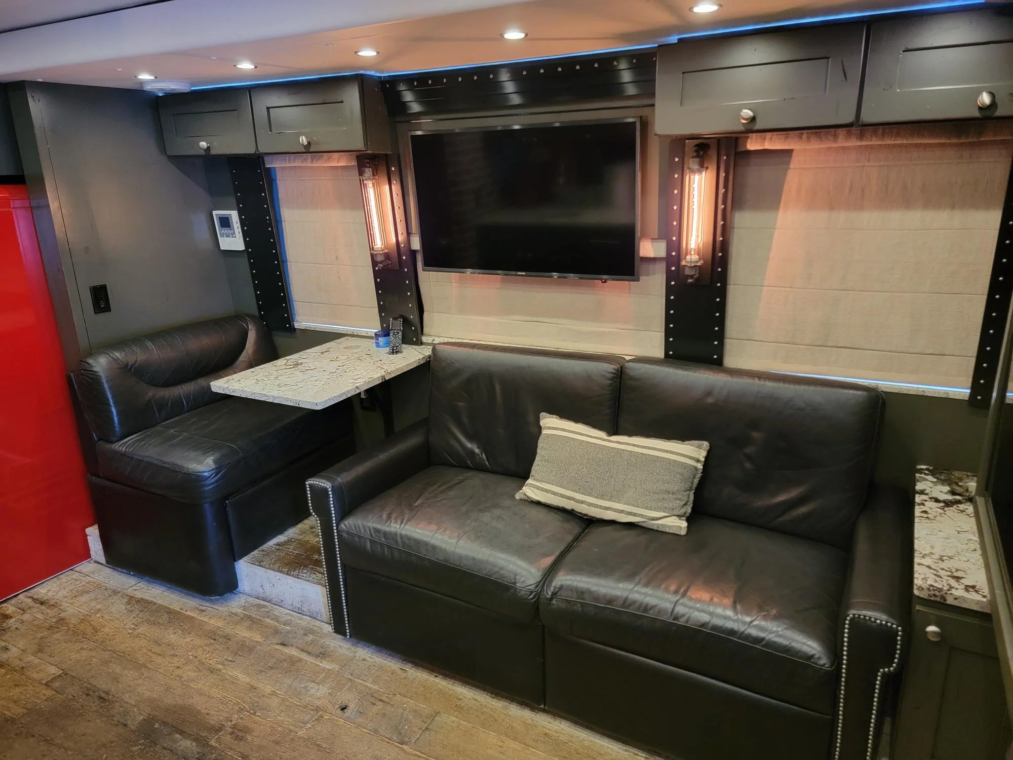 A living room area with a black leather couch, a marble-top table with a pillow on it, and a wall-mounted TV. The wall has beige blinds, decorative sconce lighting, and black accents with metal rivets. The floor is wooden, and there is a red door on 