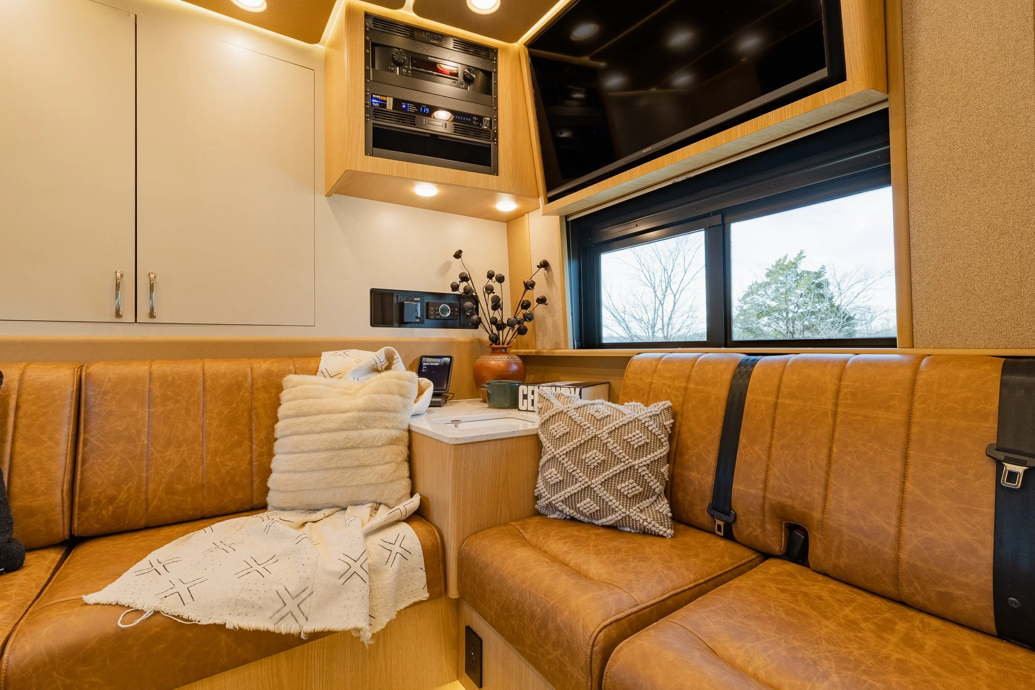 Interior of a cozy space with a window showing trees outside, a leather couch with pillows and a blanket, a small table with decorative items, a TV mounted on the wall, and built-in cabinets and electronic equipment.