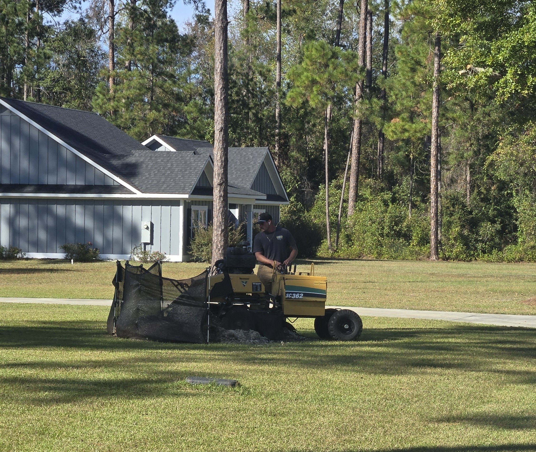 A man operating a small yellow golf course sweeper on a grassy area in front of a blue house with trees in the background on a sunny day.