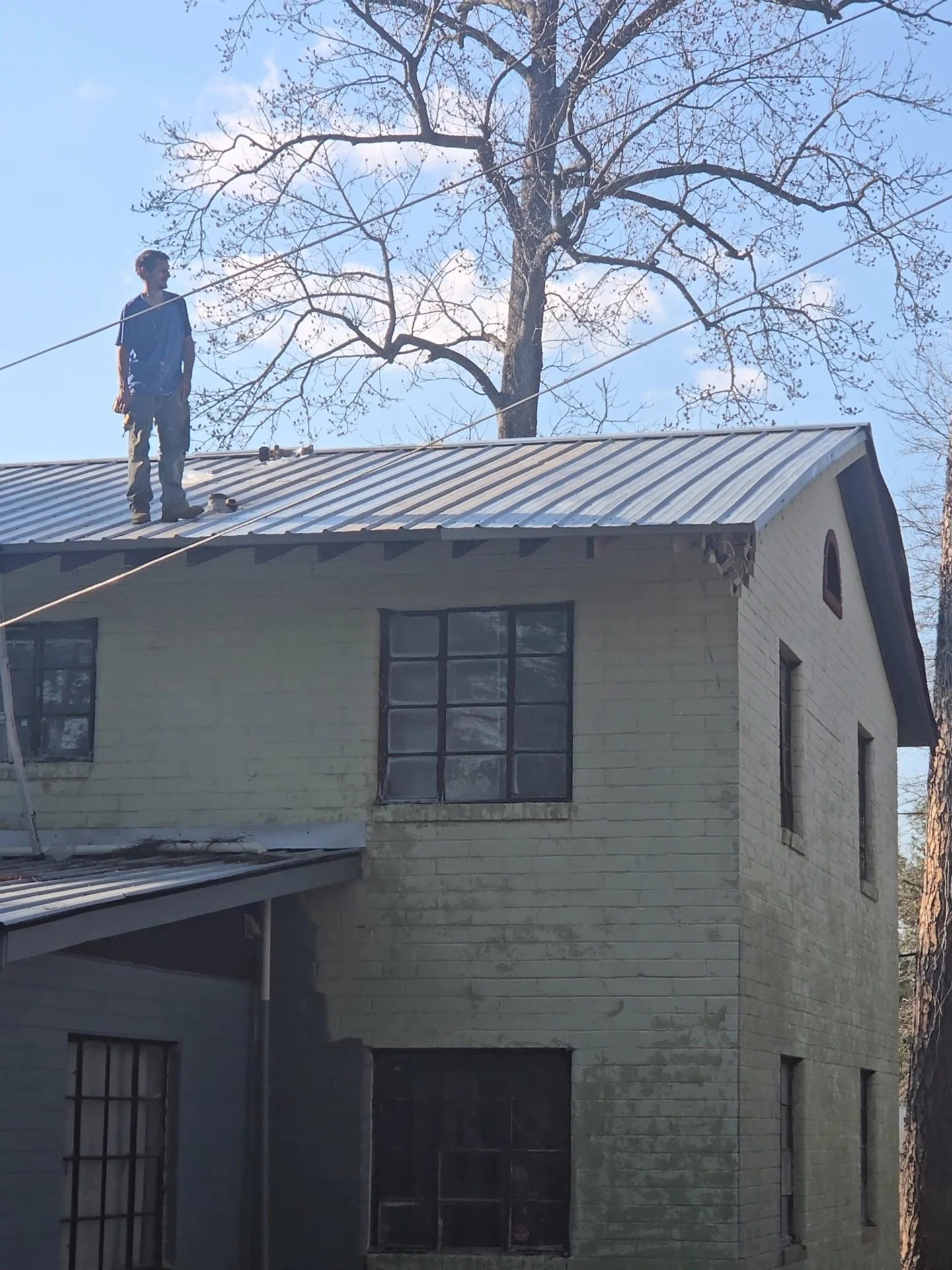A person standing on the roof of a two-story house with a metal roof, holding a safety line, with leafless trees in the background on a partly cloudy day.