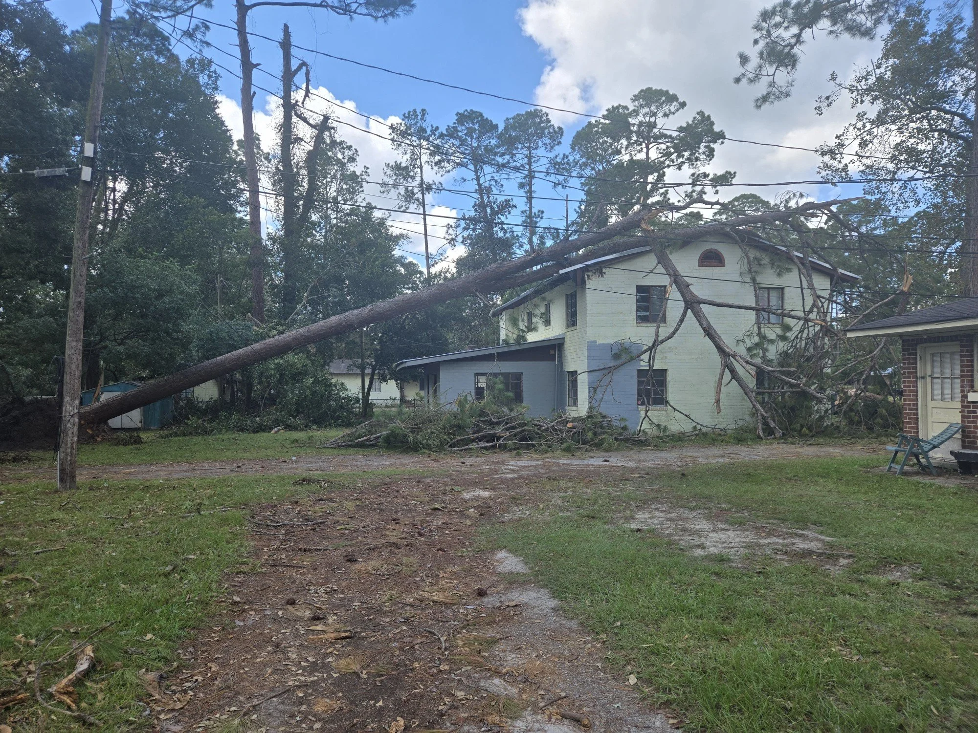 A fallen tree has collapsed onto a two-story house, causing damage to the roof and exterior walls. Debris is scattered across the yard, and the sky is partly cloudy.