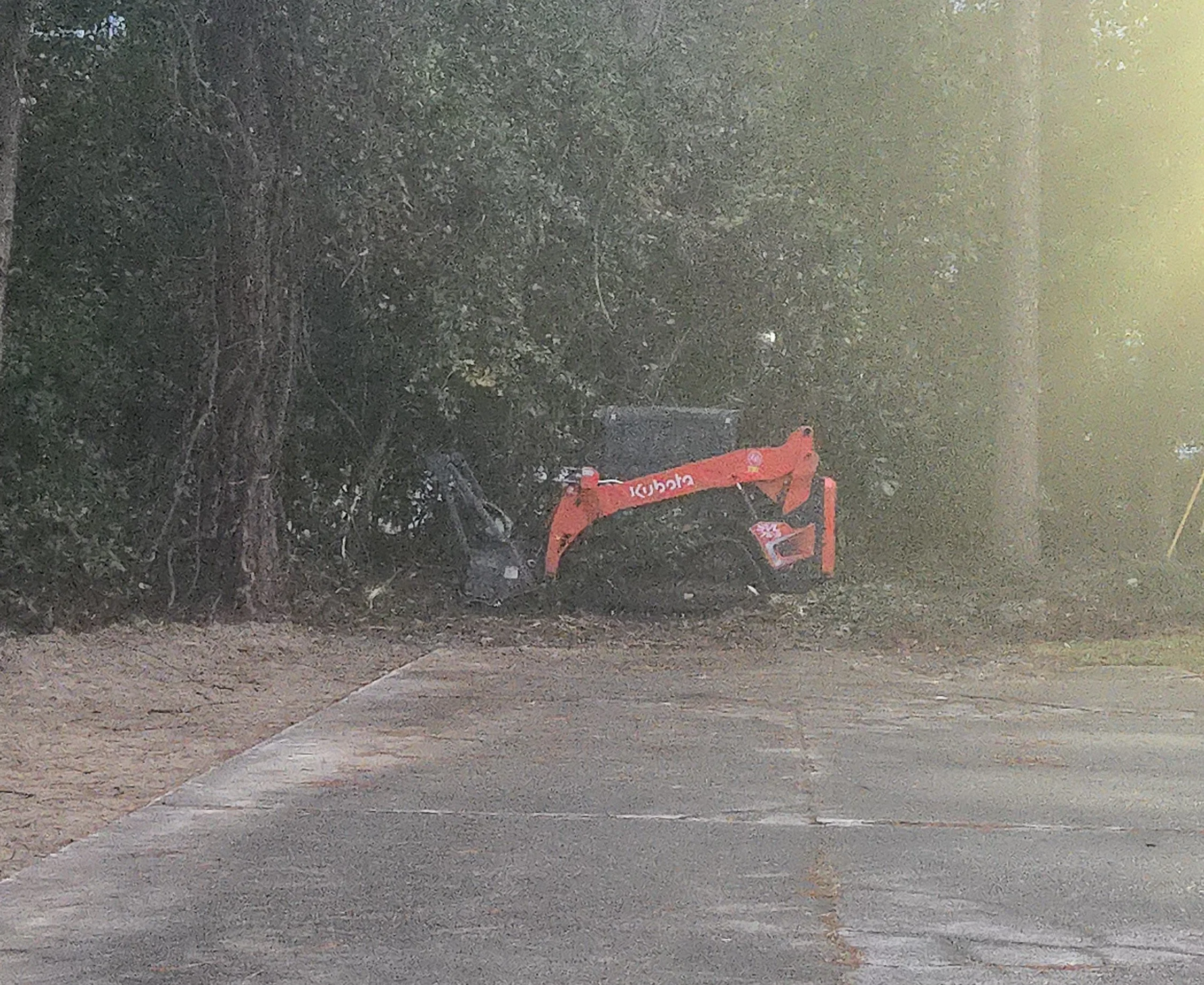 A Kubota Mini Excavator parked near a tree on a dirt path with a black object beside it, surrounded by bushes and partially obscured by a mist or fog.