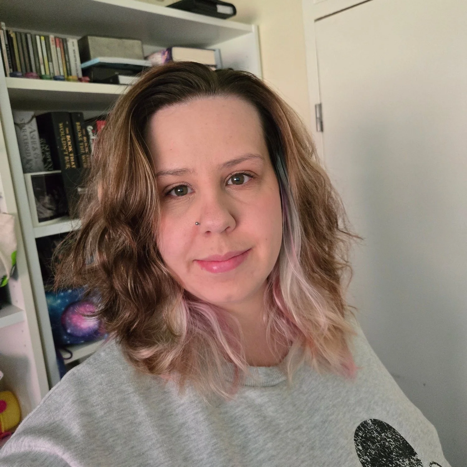 A woman with shoulder-length hair that is dark with pink tips, wearing a gray shirt with a graphic design, standing in front of a bookshelf filled with books and some decorative items.