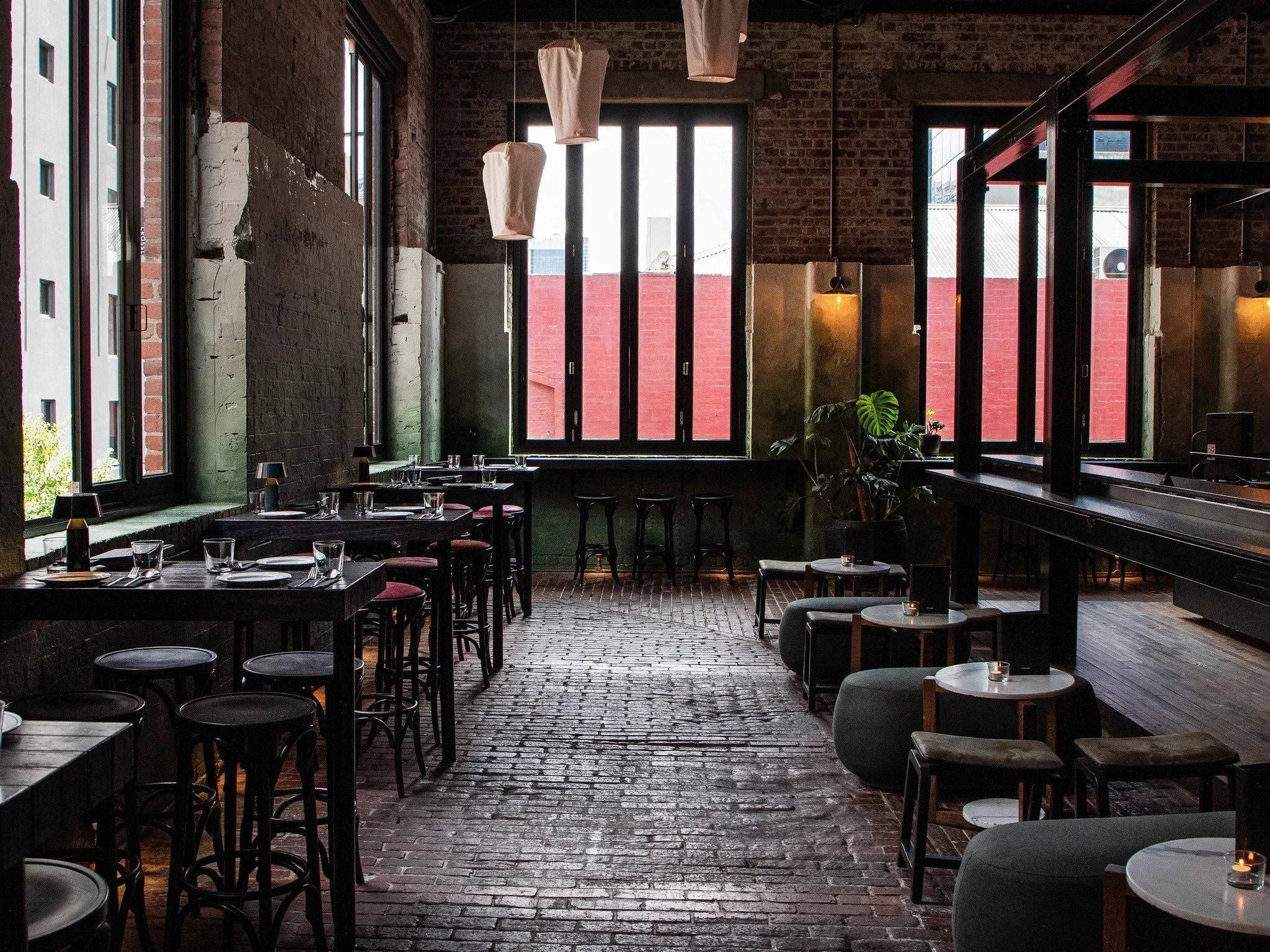 Interior of a Longsong with brick floors, large windows, and a mix of tables and seating areas, decorated with pendant lights and plants.