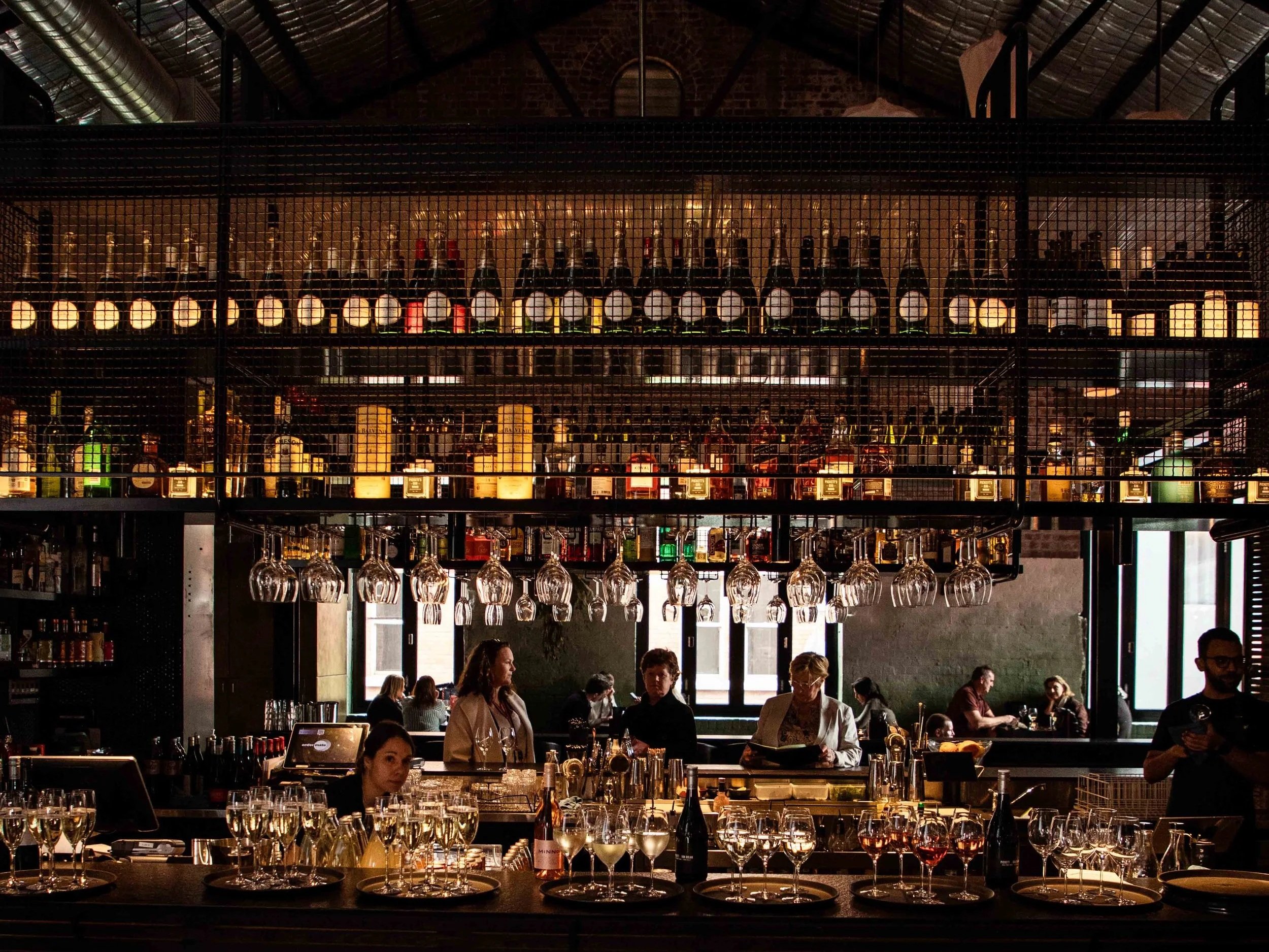 View of a well-stocked bar with hanging glassware, displayed bottles of alcohol on upper shelves, and patrons seated in the background at Longsong