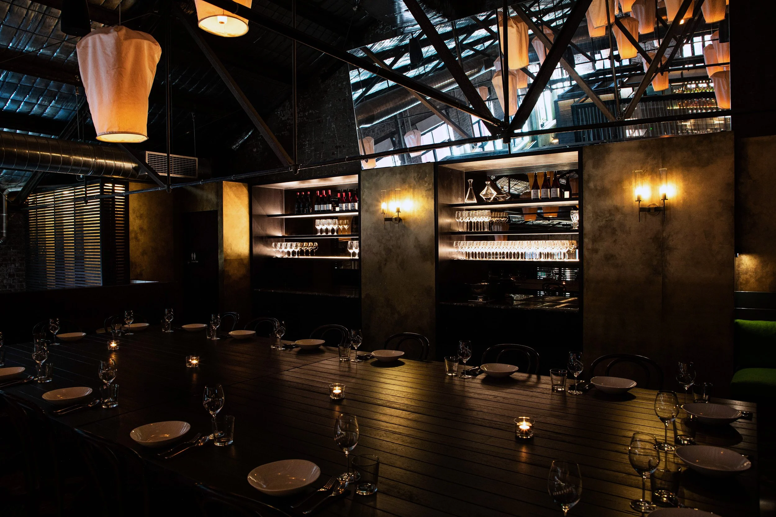 Interior of a Longsong's private dining area with a long wooden table set with glasses, plates, and candles, dim lighting, shelves with glassware and bottles on the back wall, and decorative lighting fixtures on the walls.
