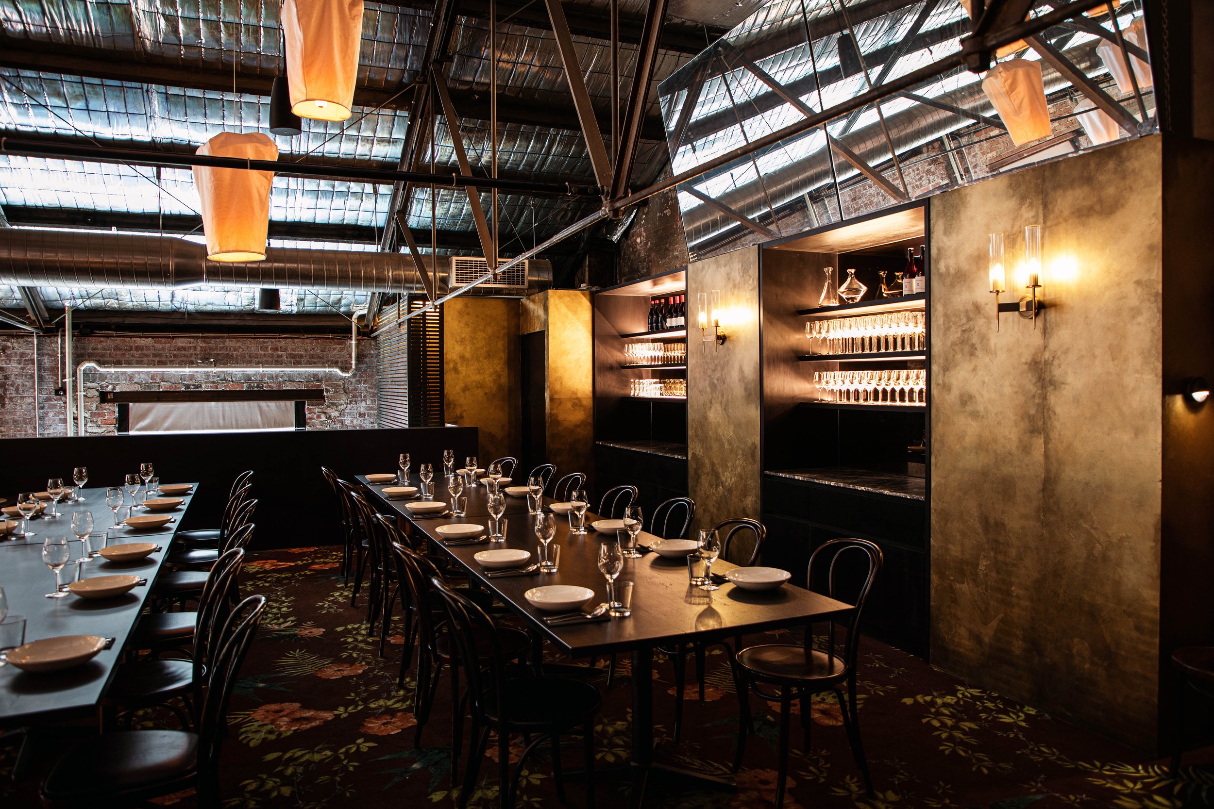 Interior of a Longsong's private dining area with a long wooden table set with glasses, plates, and candles, dim lighting, shelves with glassware and bottles on the back wall, and decorative lighting fixtures on the walls.