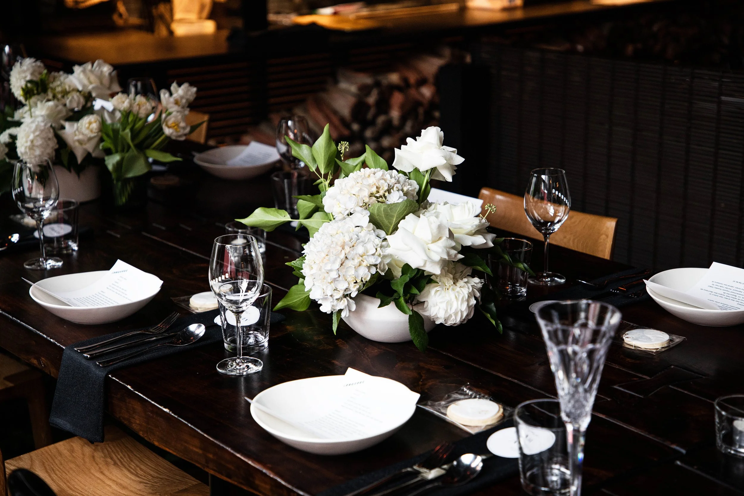 Elegant dining table set with white floral arrangement centerpiece, white bowls, wine glasses, silverware, and black napkins, in Longsong for a private event. 