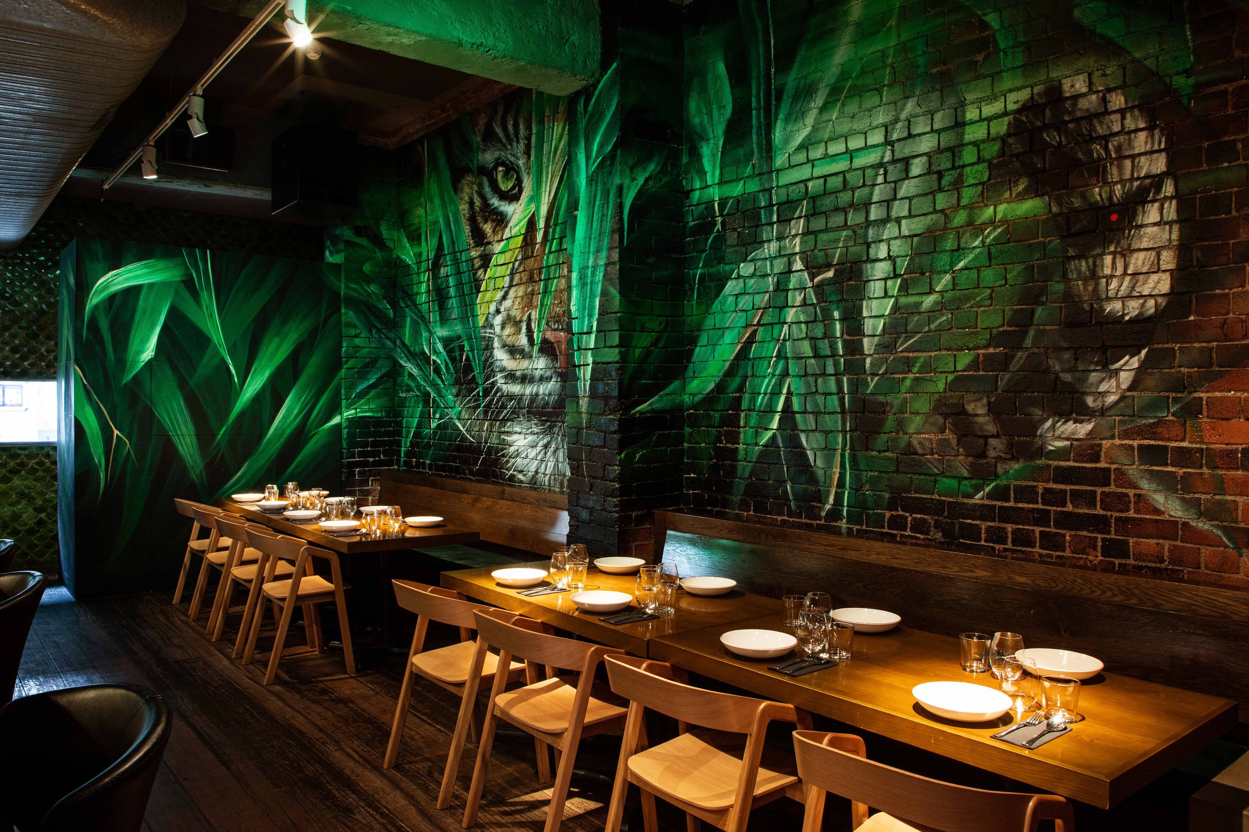 Longrain's private dining area with wooden tables set with plates and glasses, decorated with vibrant green jungle-themed mural including a tiger's face painted on brick wall.
