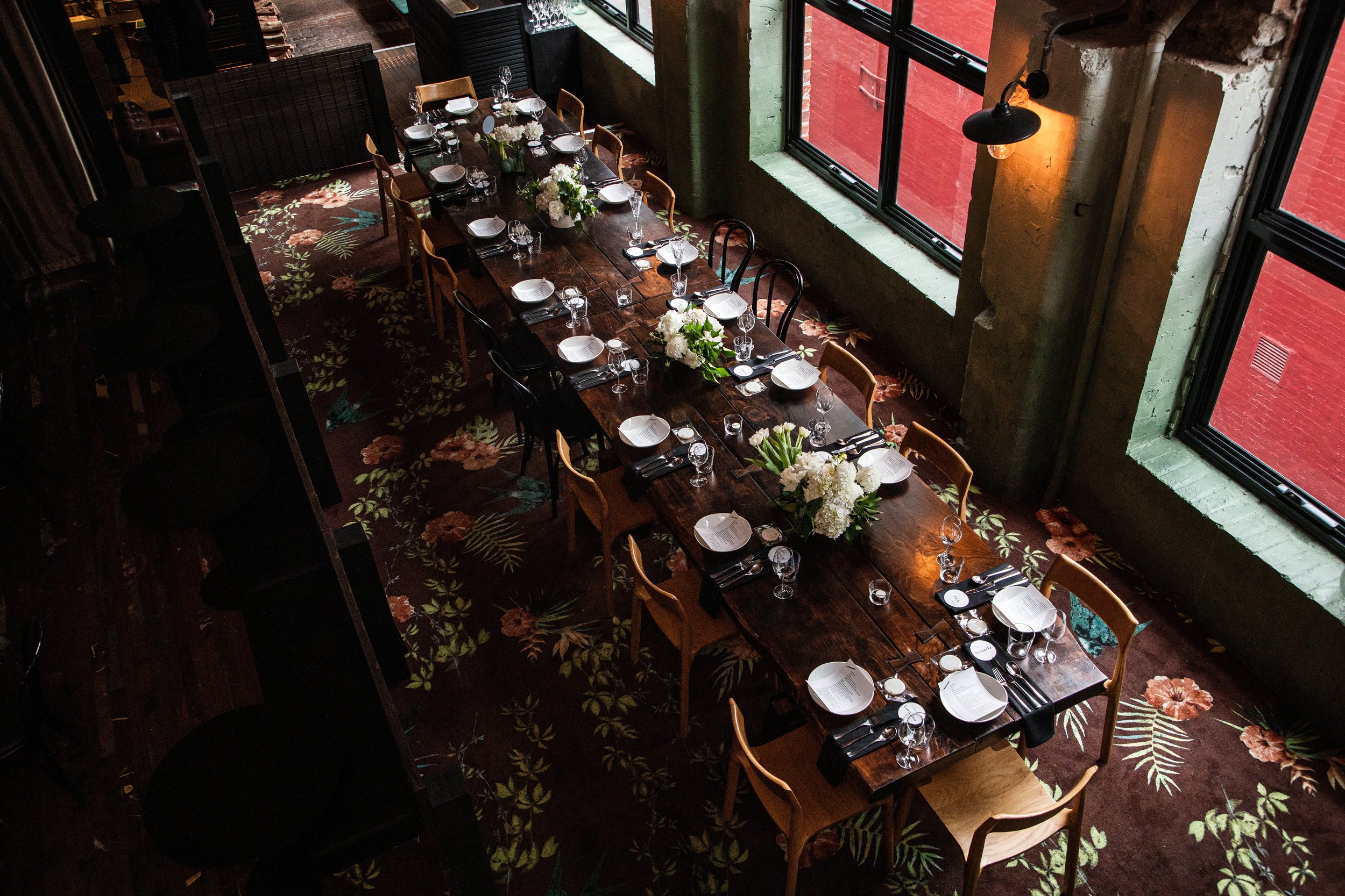 A long wooden dining table in Longsong set with white plates, silverware, and wine glasses, decorated with floral centerpieces, in a restaurant with large windows, green painted walls, and a floral patterned carpet.
