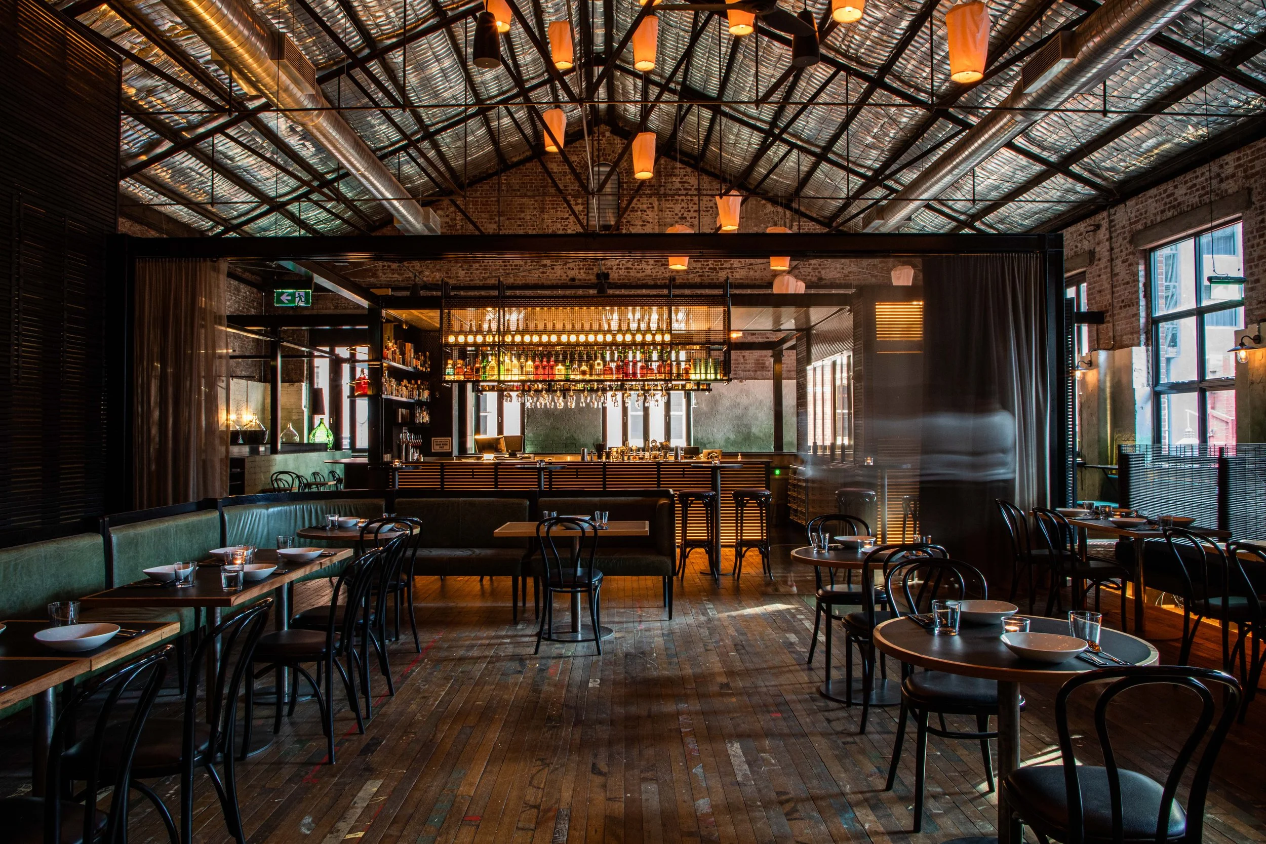 Longsong's interior with wooden floors, dark tables and chairs, a bar with bottles and hanging glasses, large windows, and warm lighting. Perfect for private events.