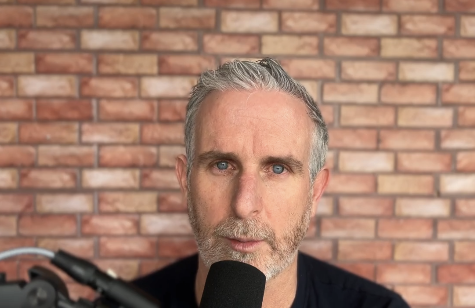 Close-up of a man with gray hair and beard speaking into a microphone against a brick wall background.
