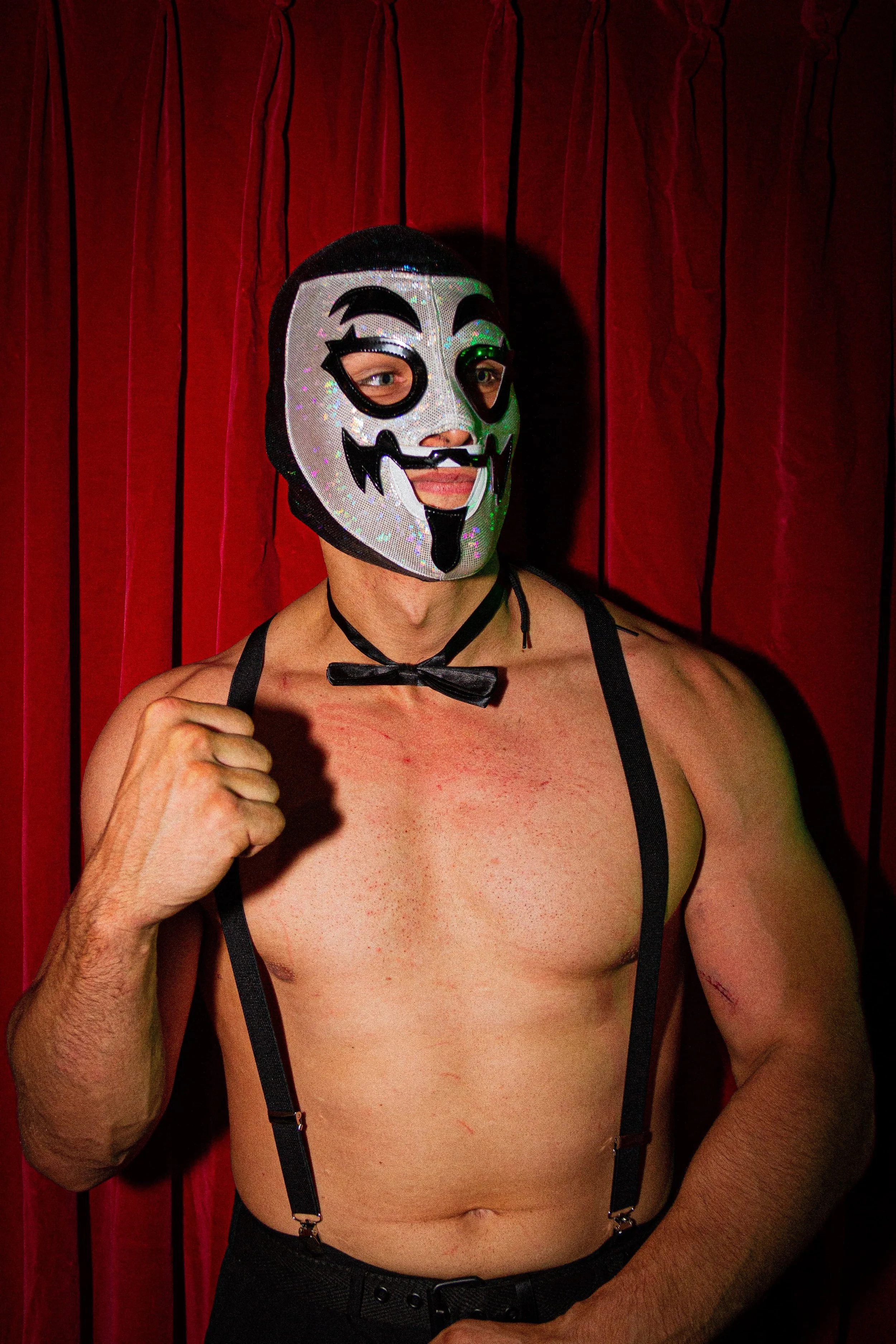 A shirtless man wearing a black and white skeleton mask, a black bow tie, suspenders, and black pants, standing in front of red curtains.