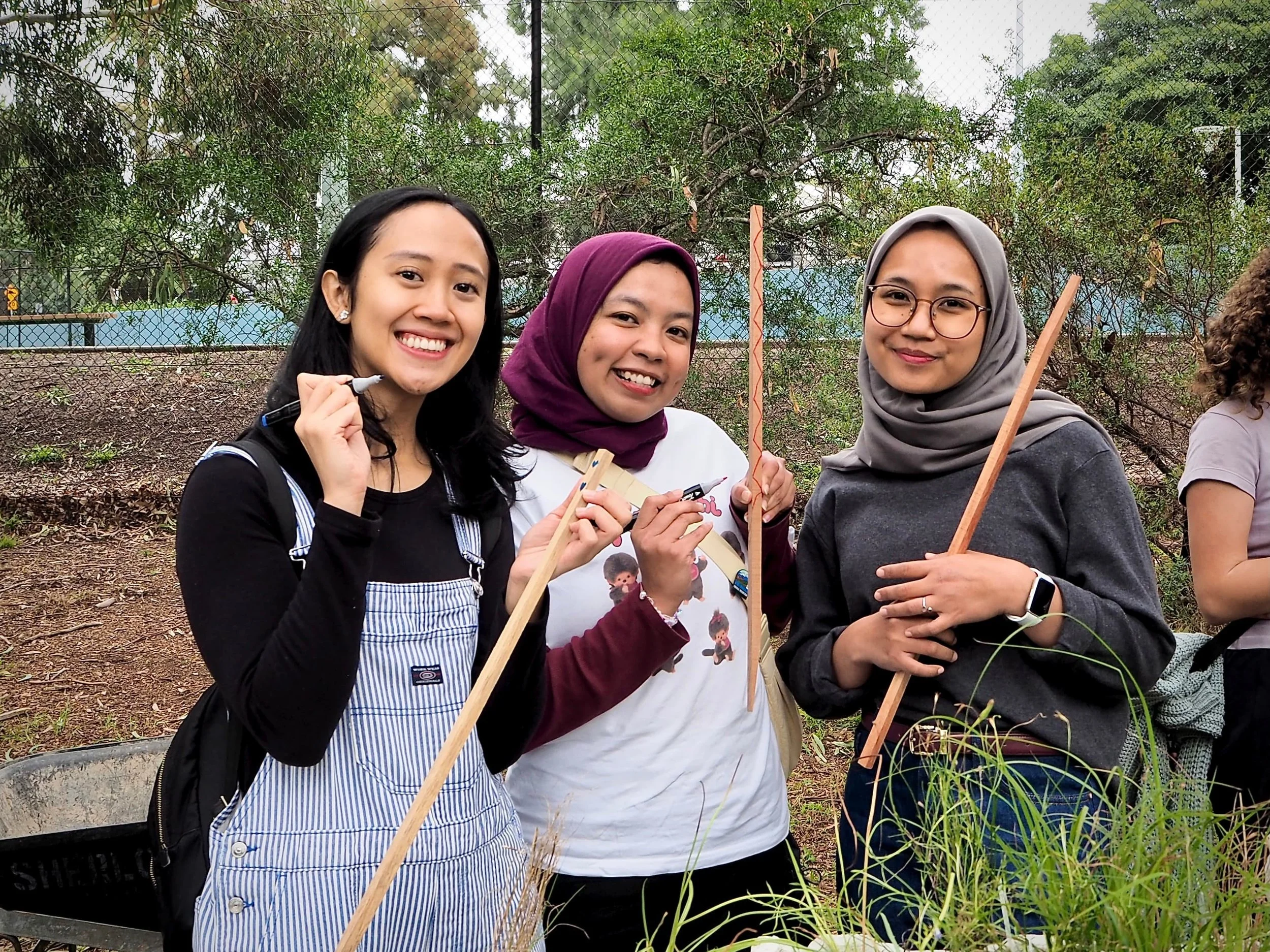 Group of three women smiling outdoors, holding sticks, with trees and fencing in the background. Three volunteers for Wattle.