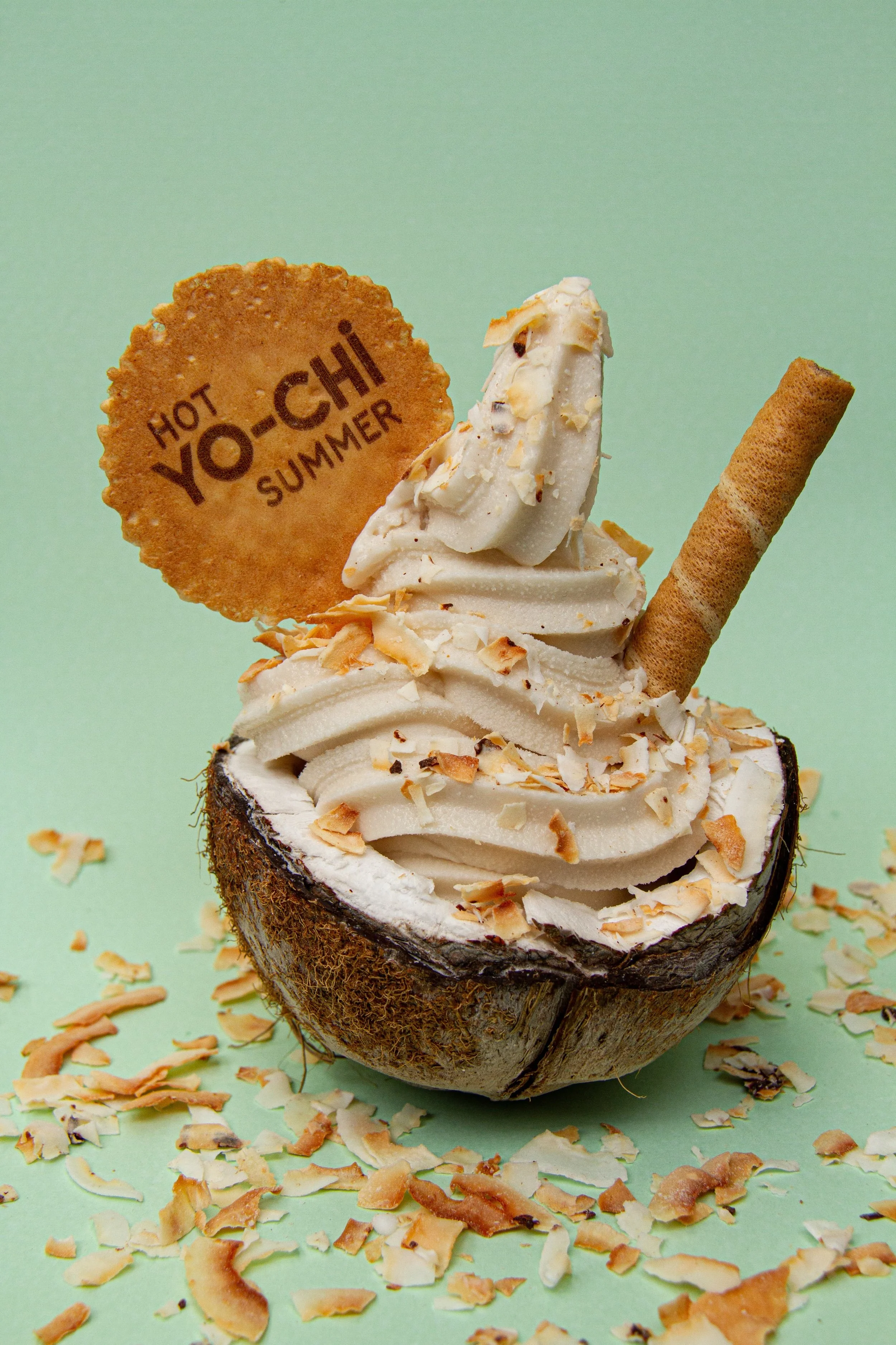 A hollowed-out coconut shell filled with soft serve ice cream topped with toasted coconut flakes, with a round cracker that reads 'Hot Yo-Chi Summer,' and a rolled wafer stick, against a light green background.