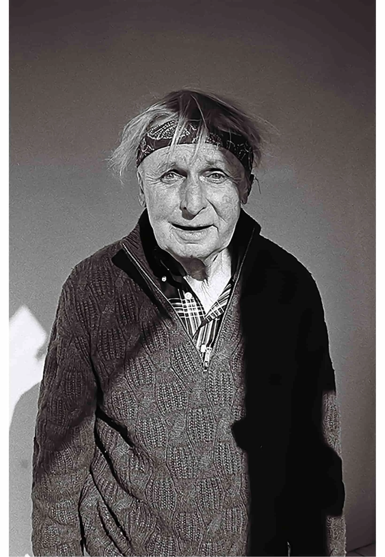 Black-and-white photo of an elderly man with light-colored hair, wearing a patterned headband, a zip-up sweater, and a shirt underneath, standing against a plain background.