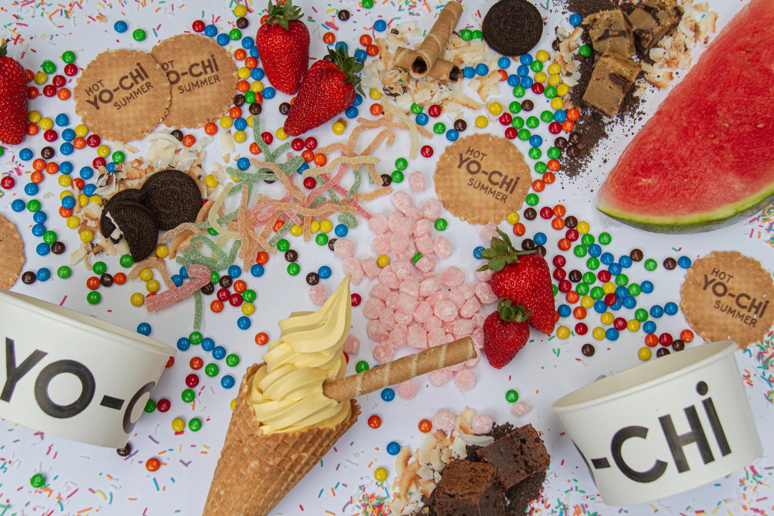 A variety of colorful candies, strawberries, watermelon slices, chocolate cookies, and whipped cream in a waffle cone. There are also some cookies, wafer rolls, and scattered sprinkles on a white surface. Two cups with 