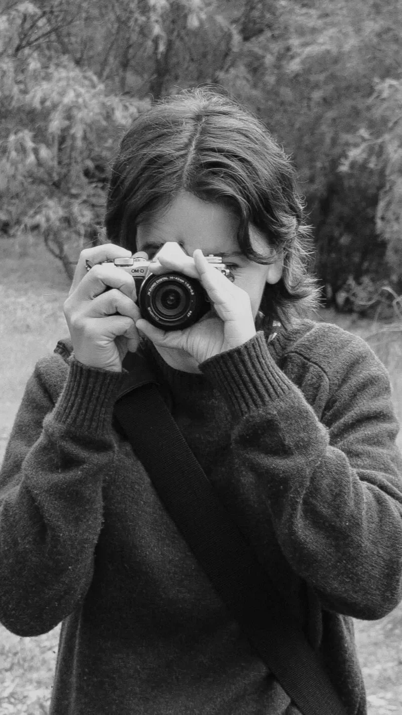A woman with short hair taking a photo with a camera outdoors, wearing a sweater and a shoulder strap, with trees in the background.