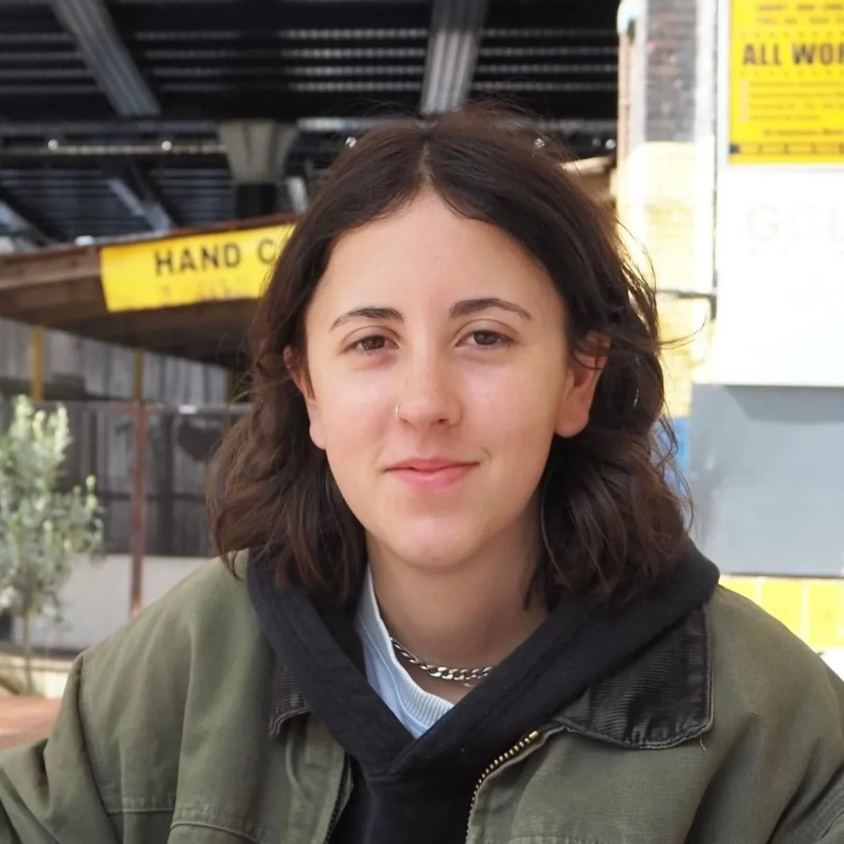 A young person with shoulder-length brown hair, light skin, wearing a silver chain necklace, a green jacket over a black hoodie, and a light blue shirt, sitting outdoors near a metal structure with yellow signs reading 'Hand C...' and another yellow sign in background.