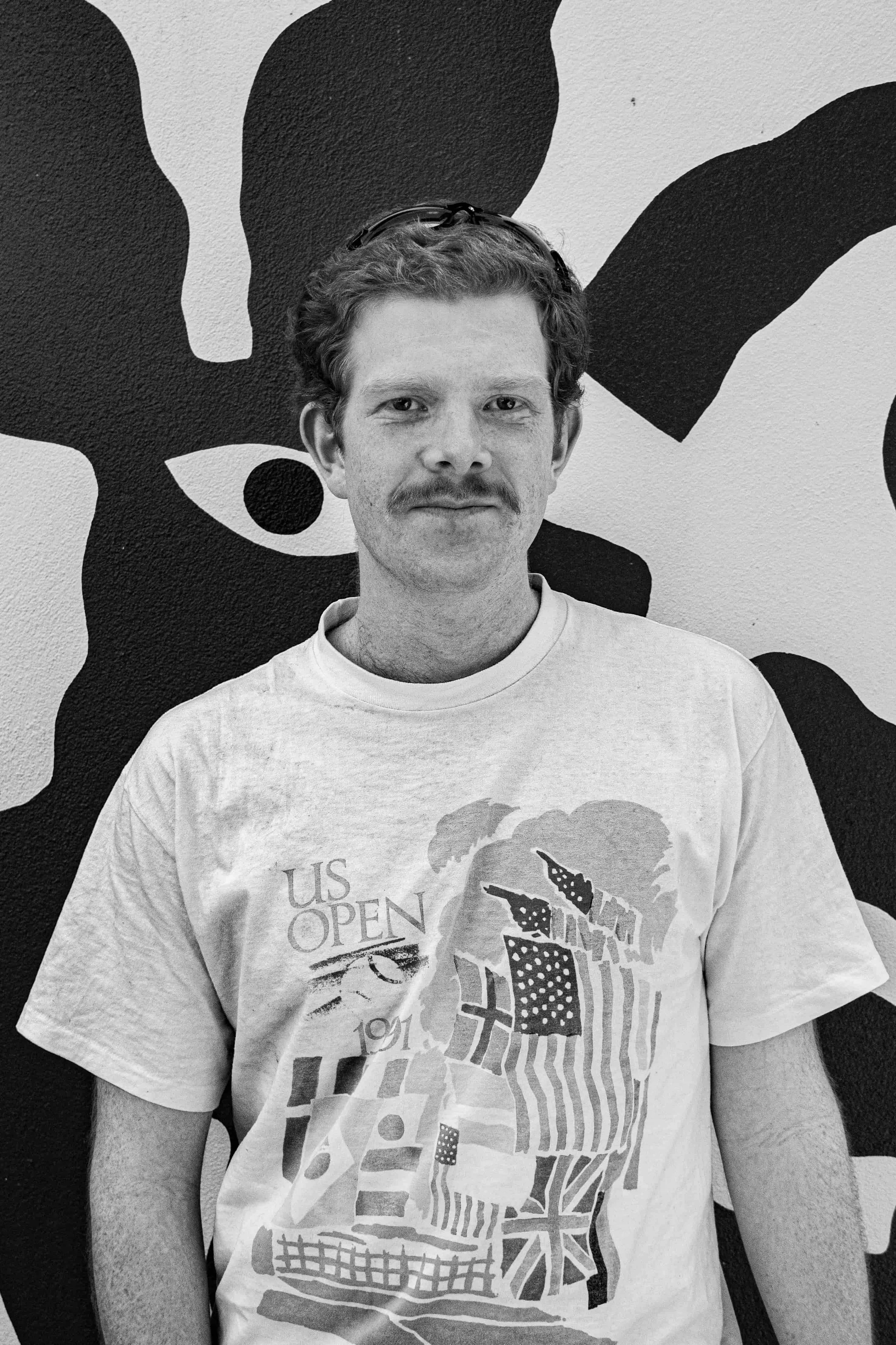 A man with curly hair, a mustache, and glasses on his head wearing a t-shirt with a graphic design and text, standing in front of a black and white abstract wall art.