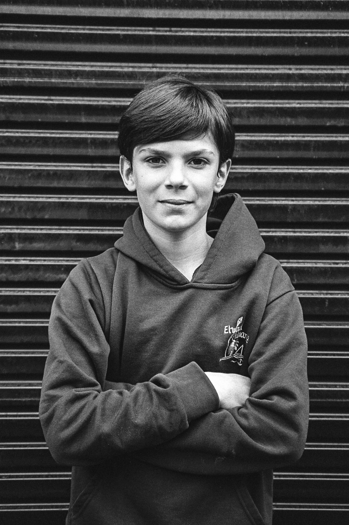 A black and white photo of a young boy with short hair, wearing a hoodie with a logo on it, standing in front of a striped metal background. He is crossing his arms and looking directly at the camera with a slight smile.
