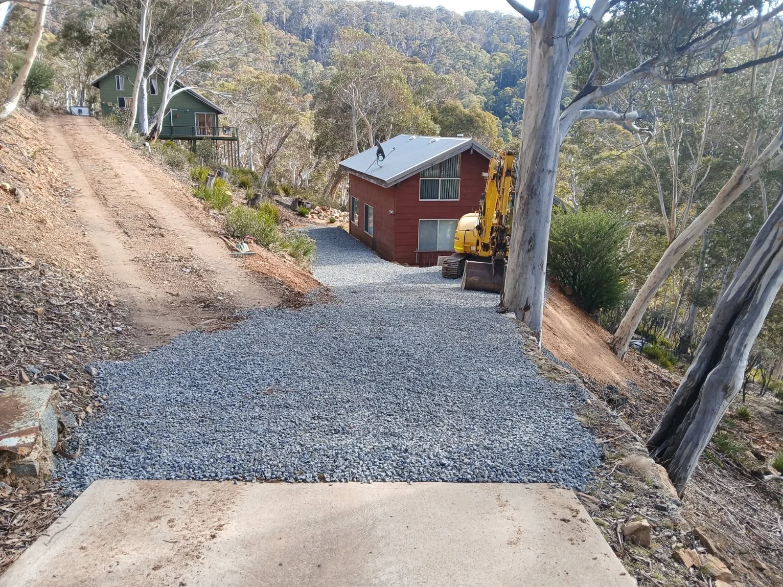 Residential construction site showing gravel pathway leading to red house, with construction equipment nearby and wooded hillside in the background.